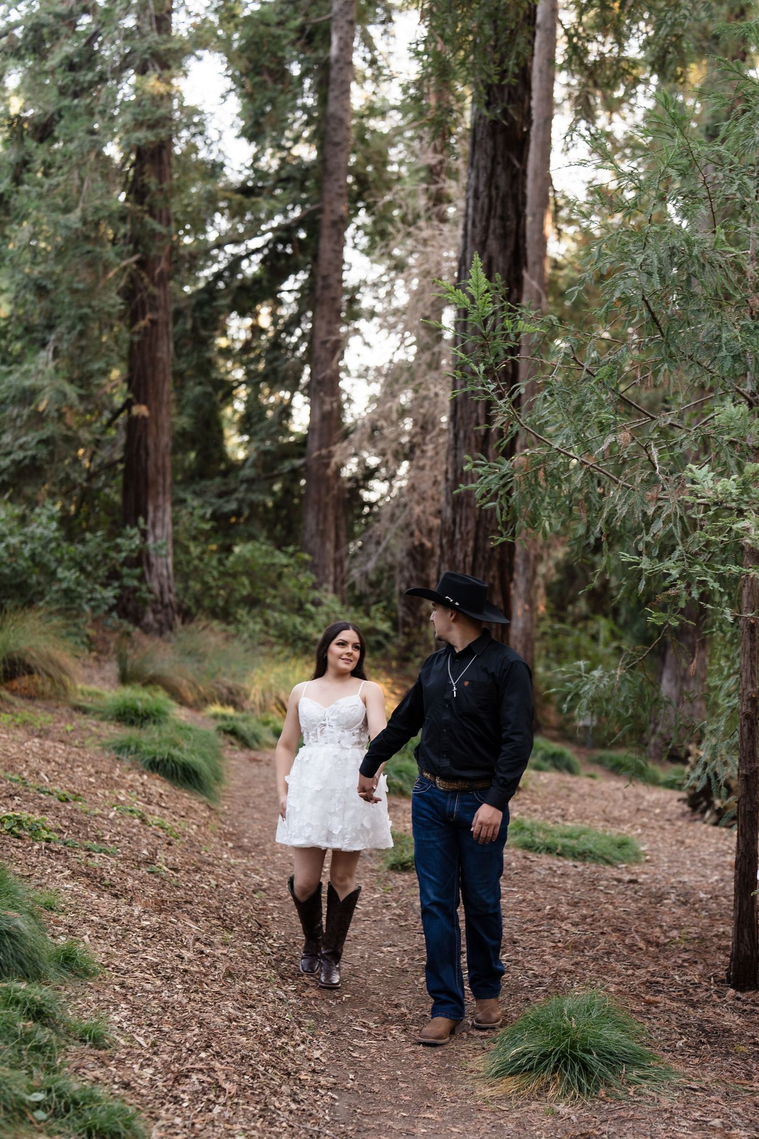 Couple walking hand-in-hand through a redwood forest, woman in white dress and cowboy boots, man in black cowboy hat.