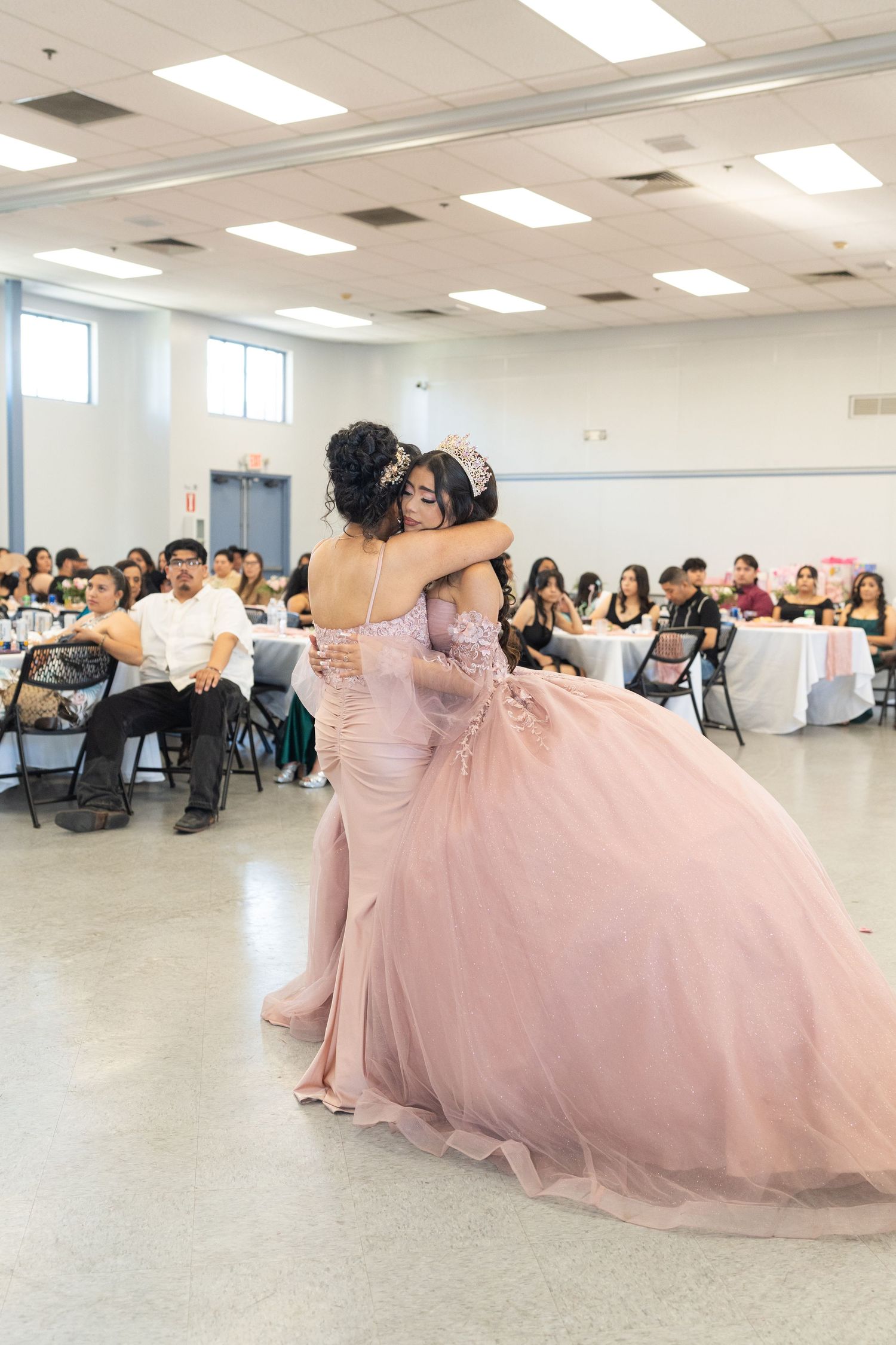 Two women in pink quinceañera gowns share an emotional hug on the dance floor at a reception.