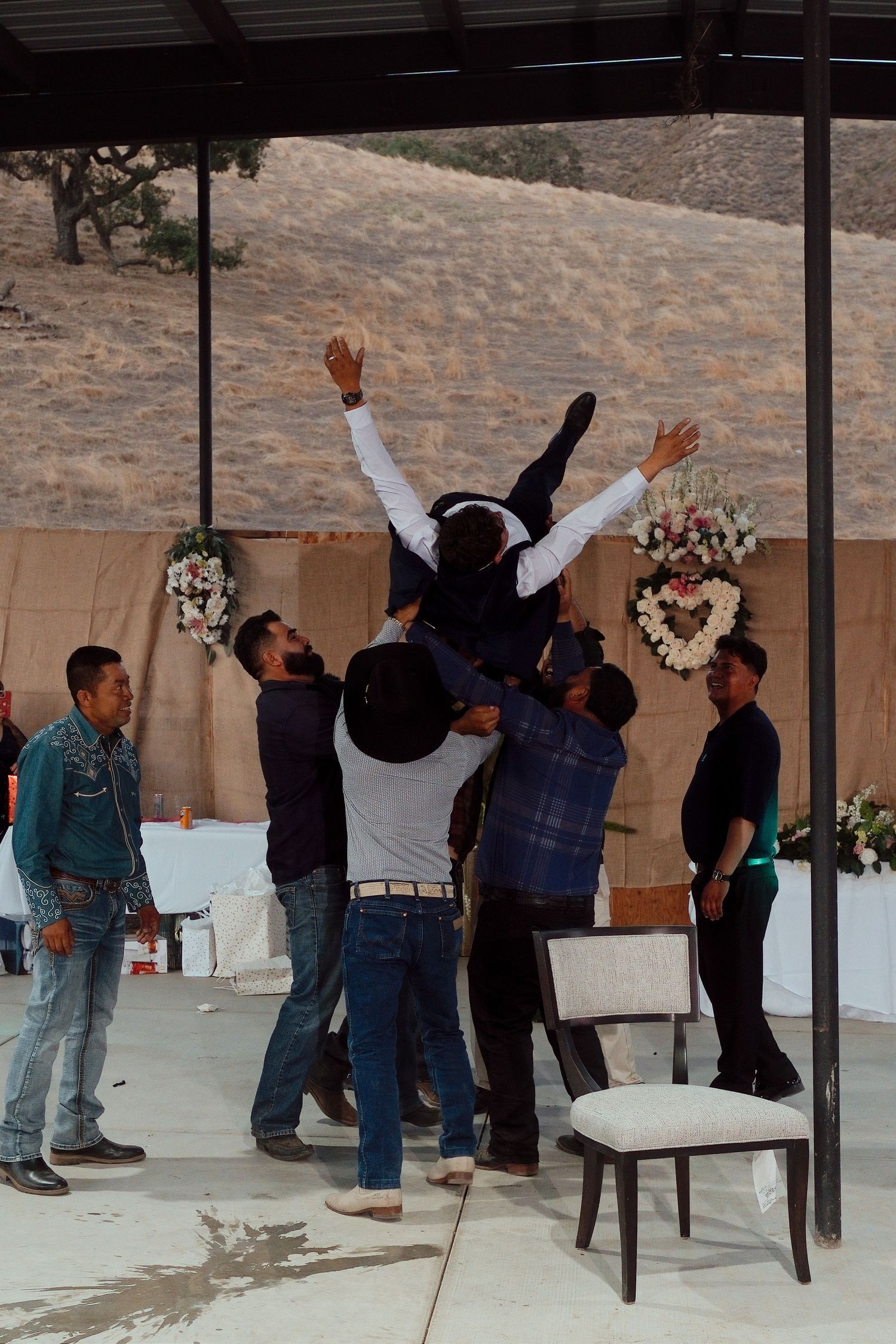 Groomsmen lifting the groom into the air in celebration at an outdoor wedding venue with floral arrangements.