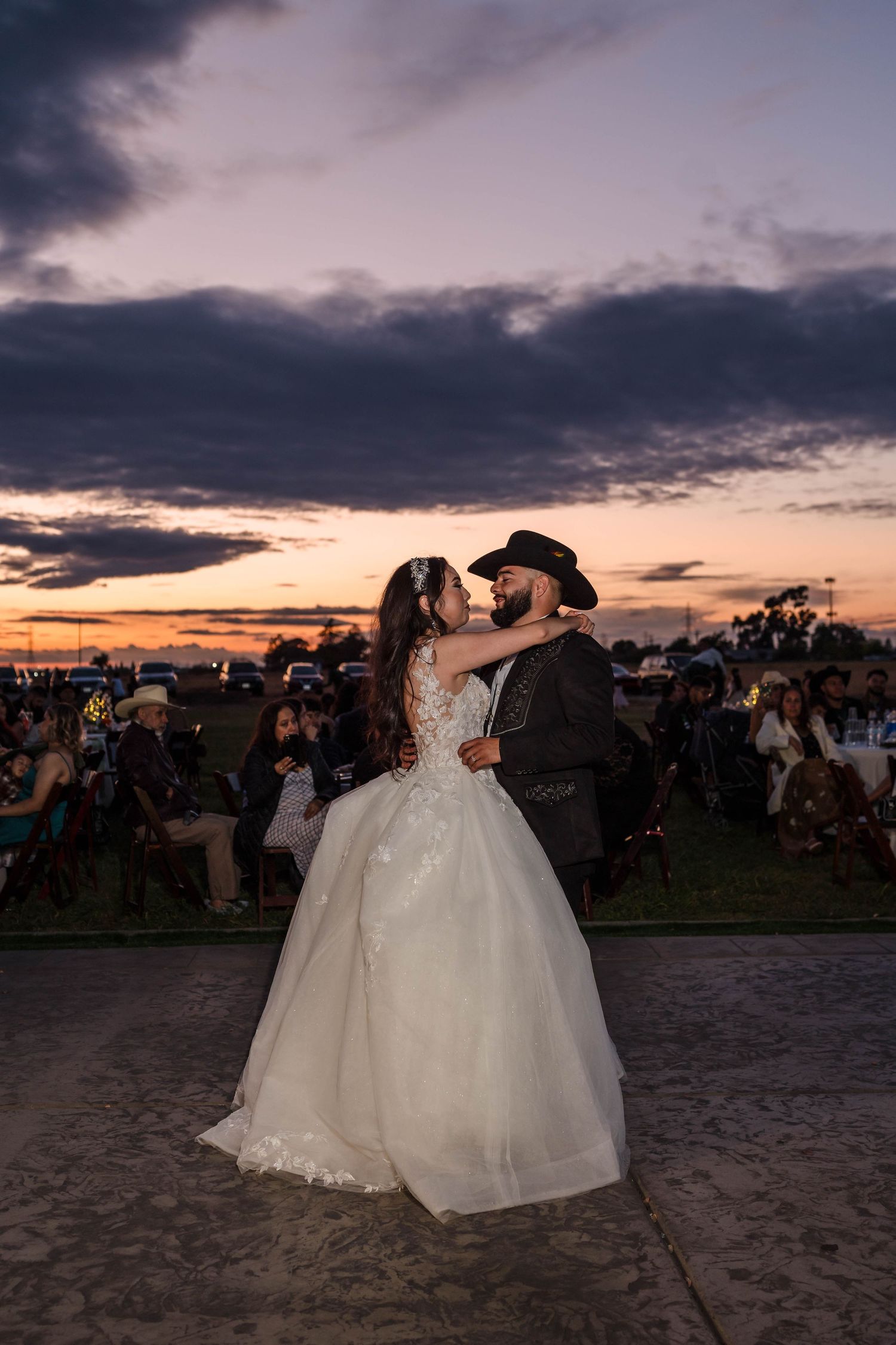 Bride and groom share first dance at sunset wedding, groom wearing cowboy hat, dramatic purple sky backdrop.