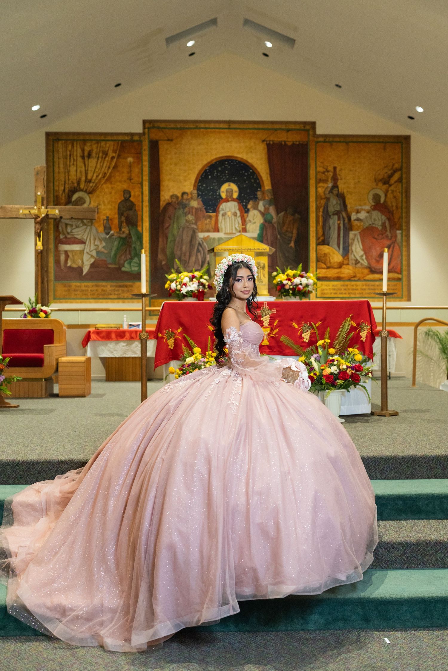Quinceañera in pink ball gown poses at church altar with religious artwork and floral arrangements.