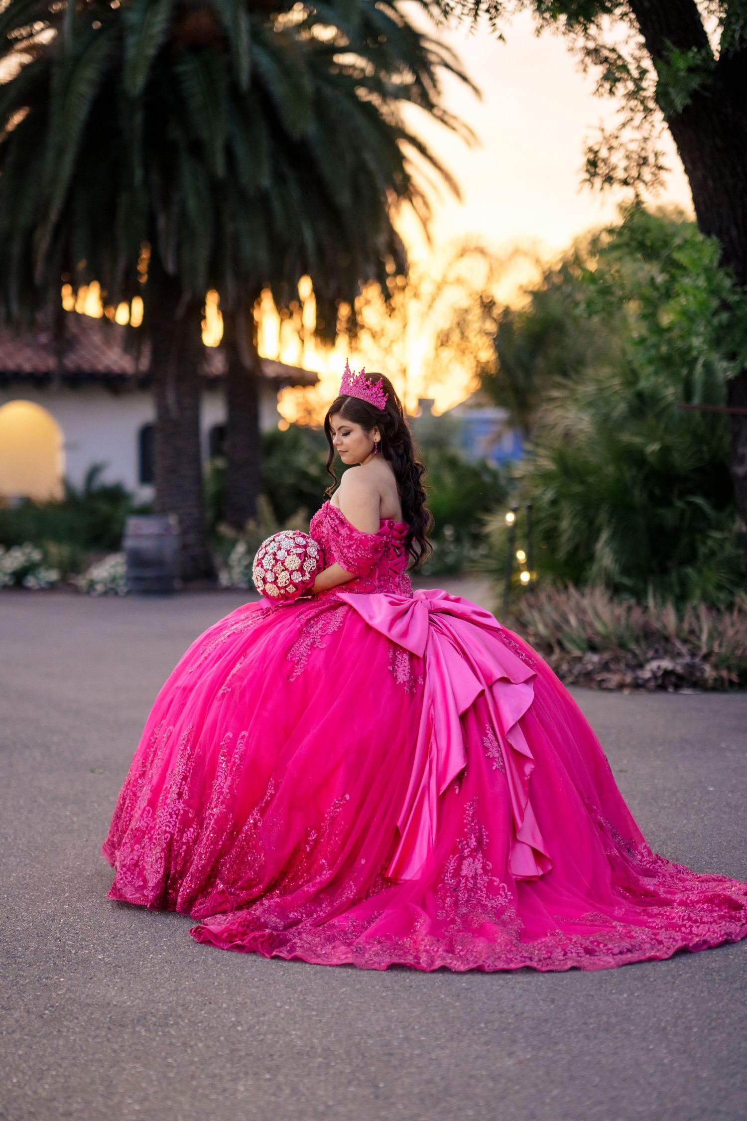 Quinceañera in hot pink ball gown with large bow and crown poses at sunset in garden.