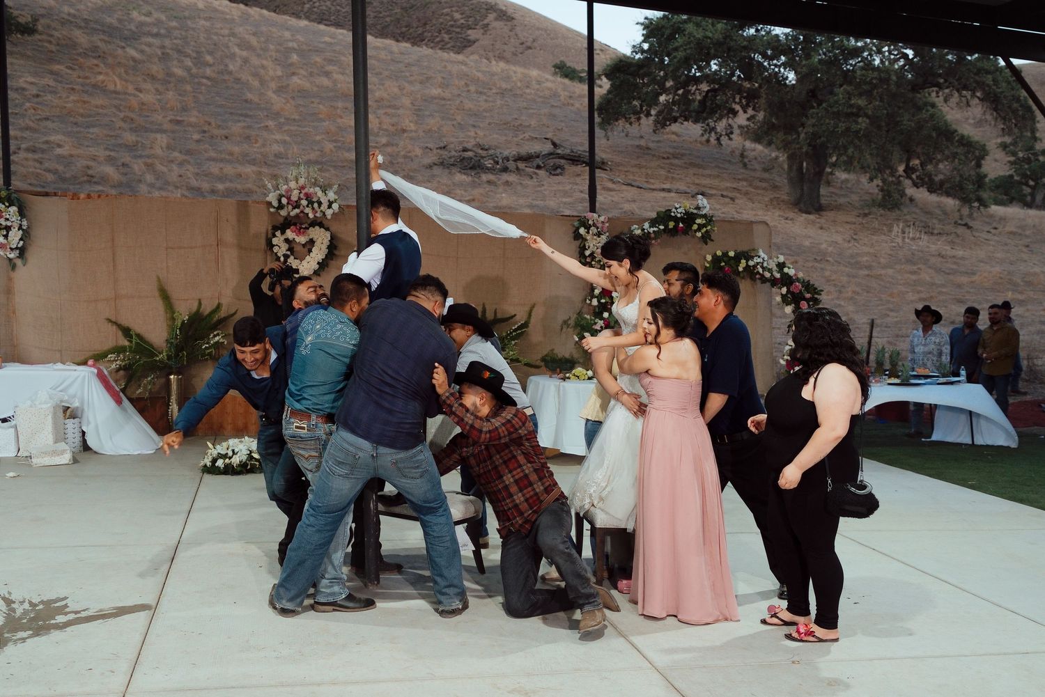 Wedding guests gather around bride and groom at outdoor reception venue with floral arch decorations.