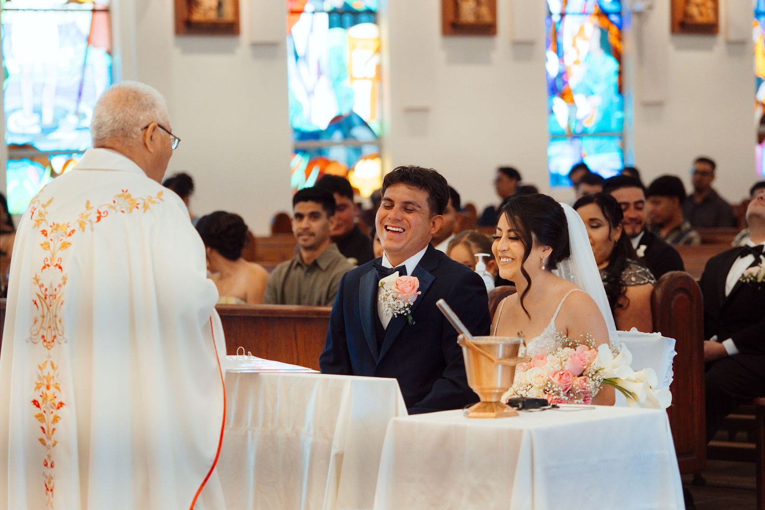 Joyful couple exchanging vows at a church wedding ceremony with a priest in white vestments and stained glass windows.