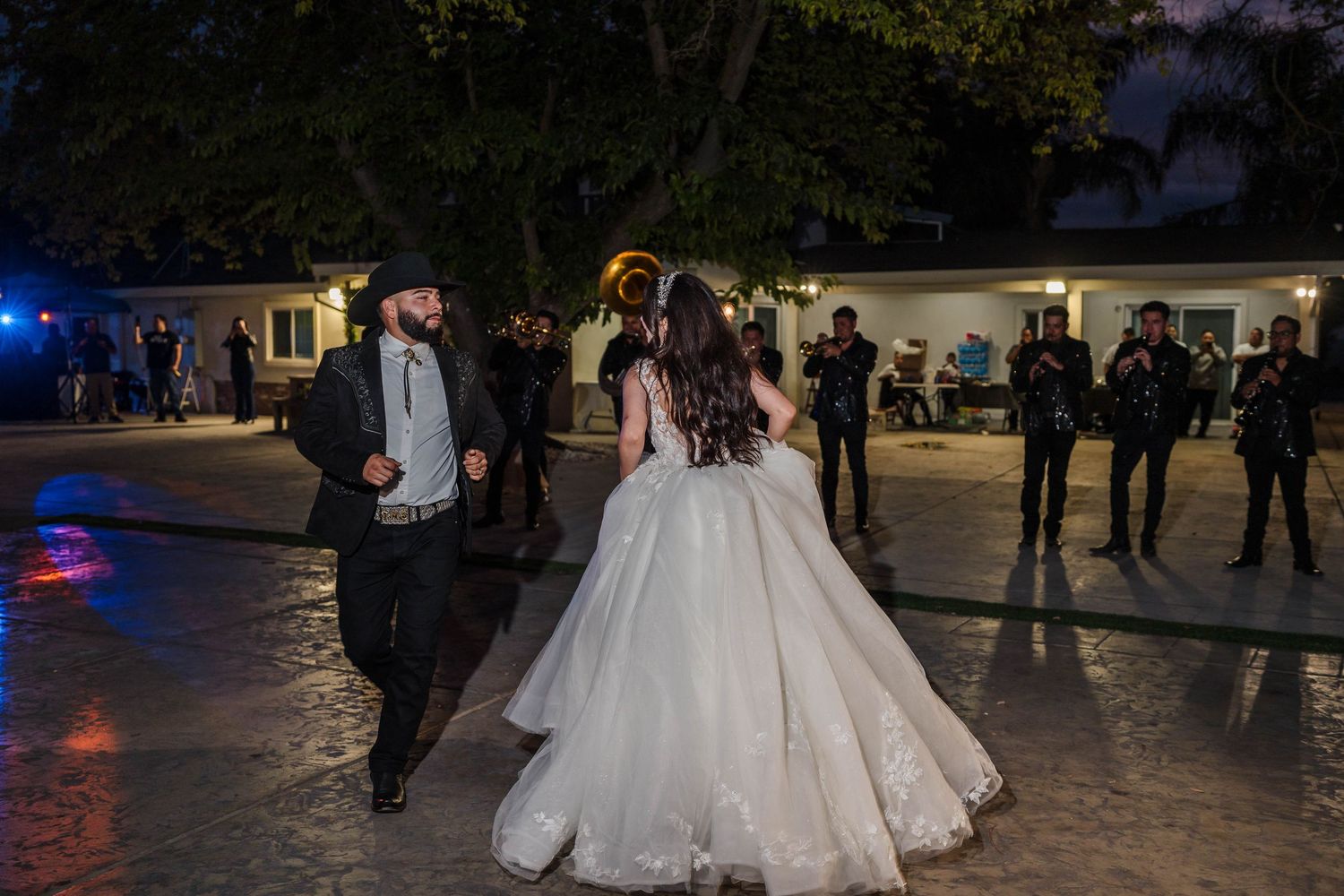 Couple sharing first dance at outdoor wedding reception at night, bride in white ballgown, mariachi band performing nearby.