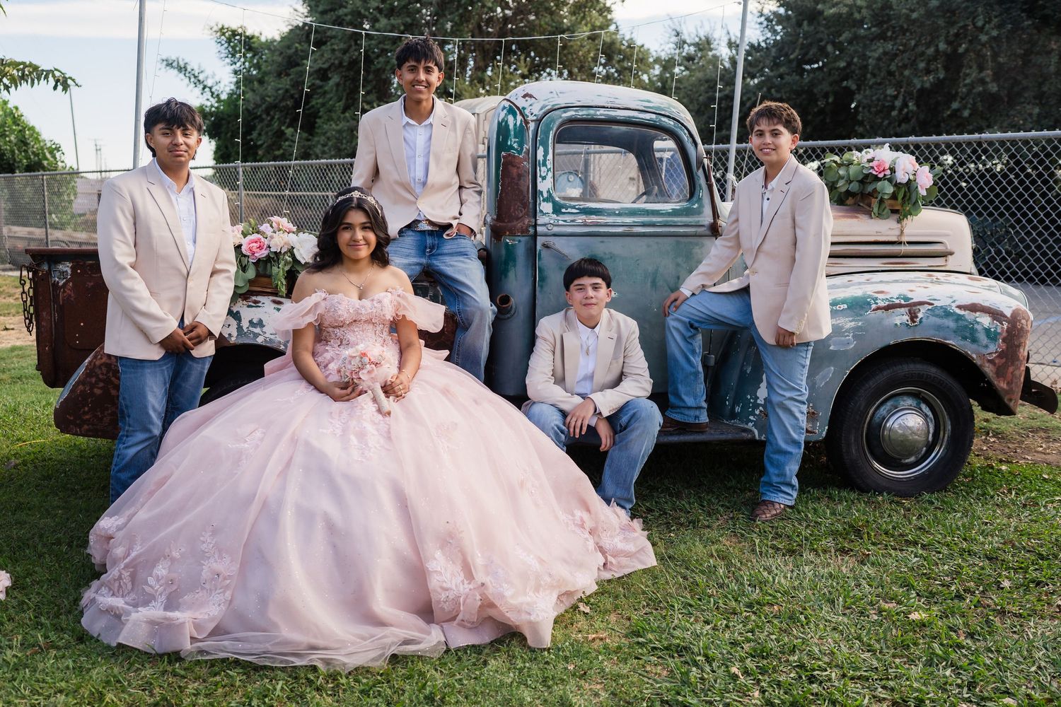 Quinceañera girl in pink gown poses with four young men beside vintage blue truck with flowers.