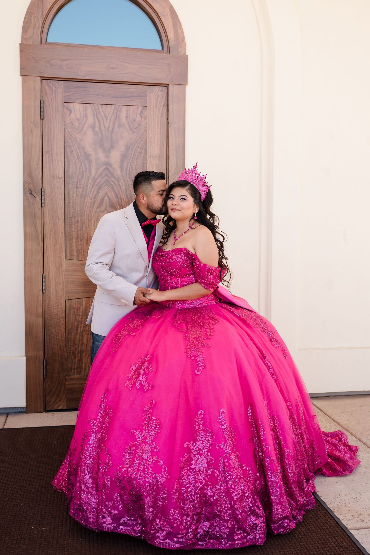 Young woman in a vibrant pink quinceañera ballgown with crown poses with a man in a white blazer by a wooden door.
