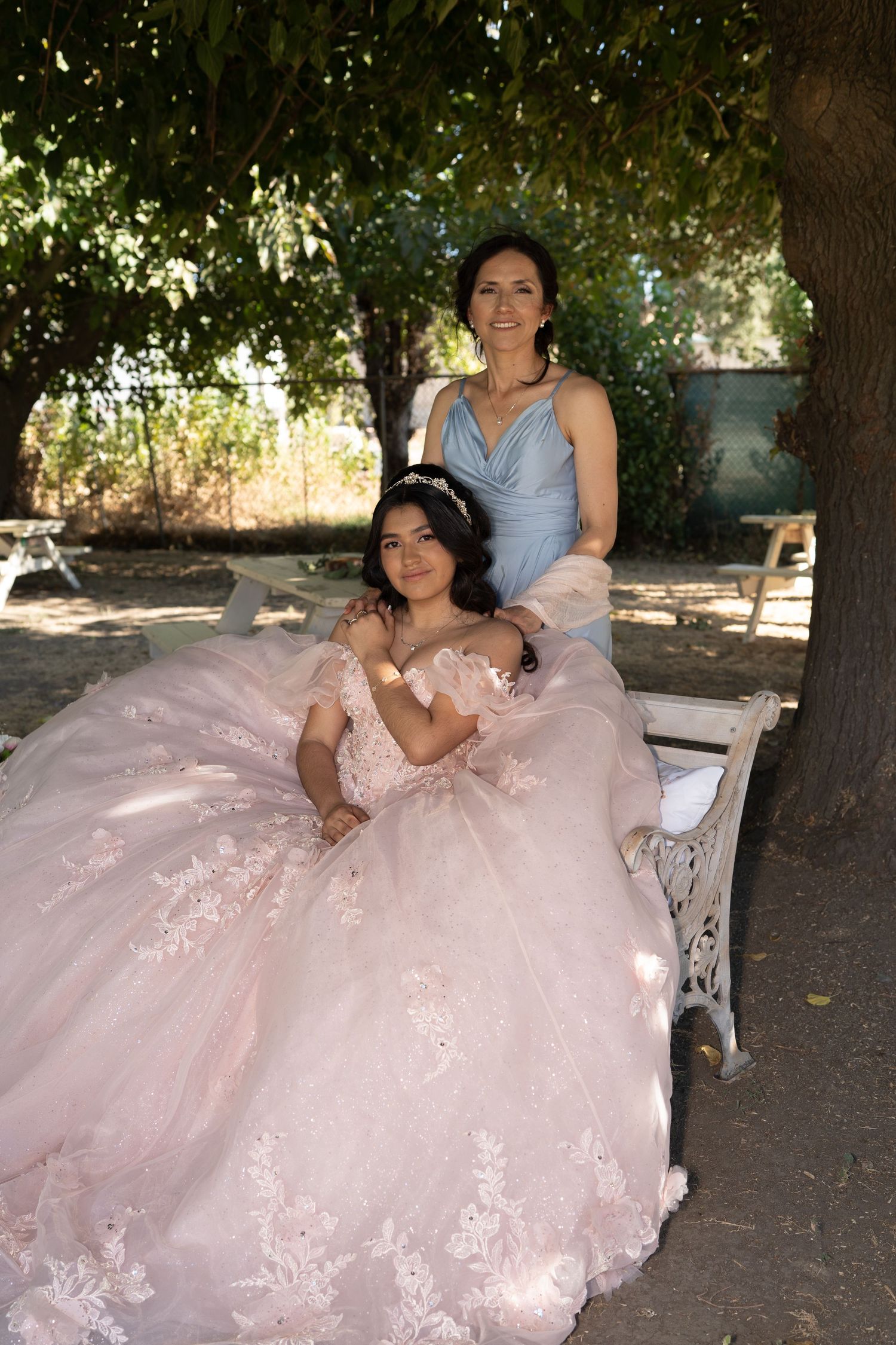A young girl in a pink quinceañera gown sits on a bench with a woman in blue dress beside her outdoors.