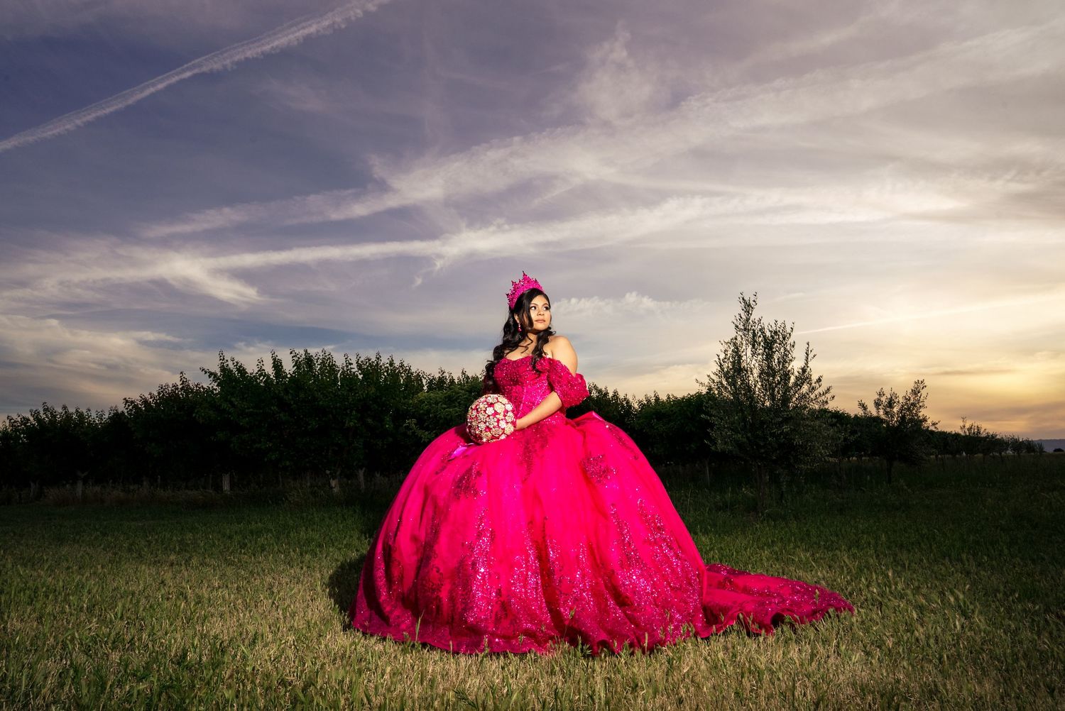 Quinceañera in dramatic fuchsia ball gown holds bouquet while posing in field at golden sunset.