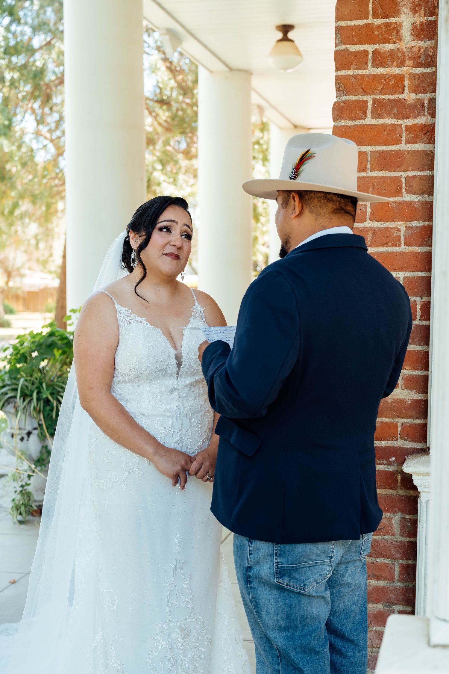 Bride and groom hold hands during emotional first look on a sunlit porch with white columns.