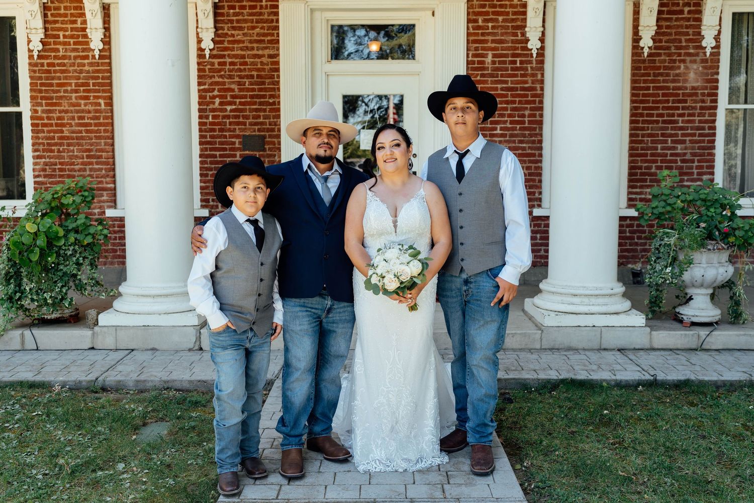 Wedding party of four in cowboy hats and jeans posing in front of a brick building.