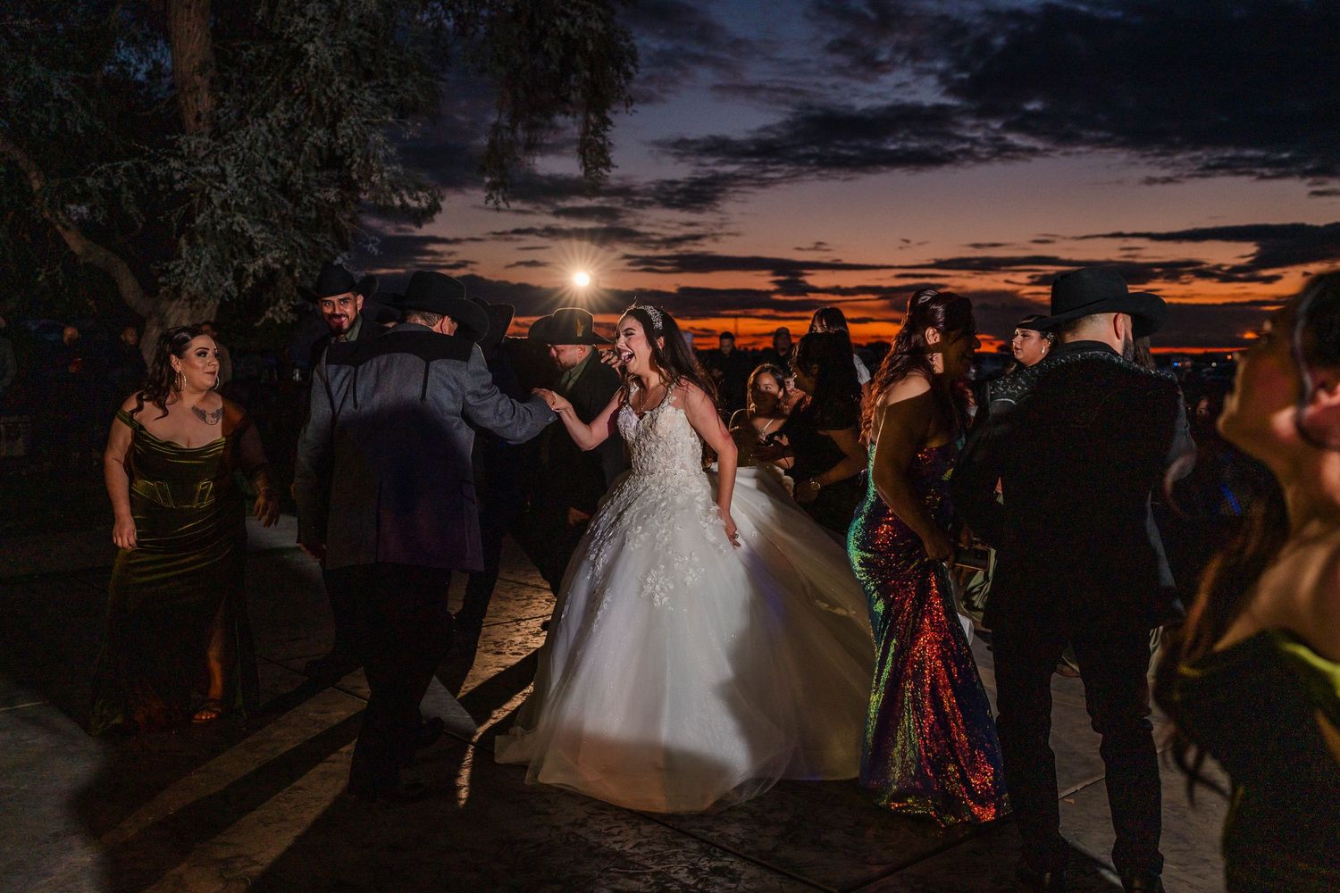 Bride in white ball gown dances joyfully with wedding guests outdoors under a stunning colorful sunset sky.