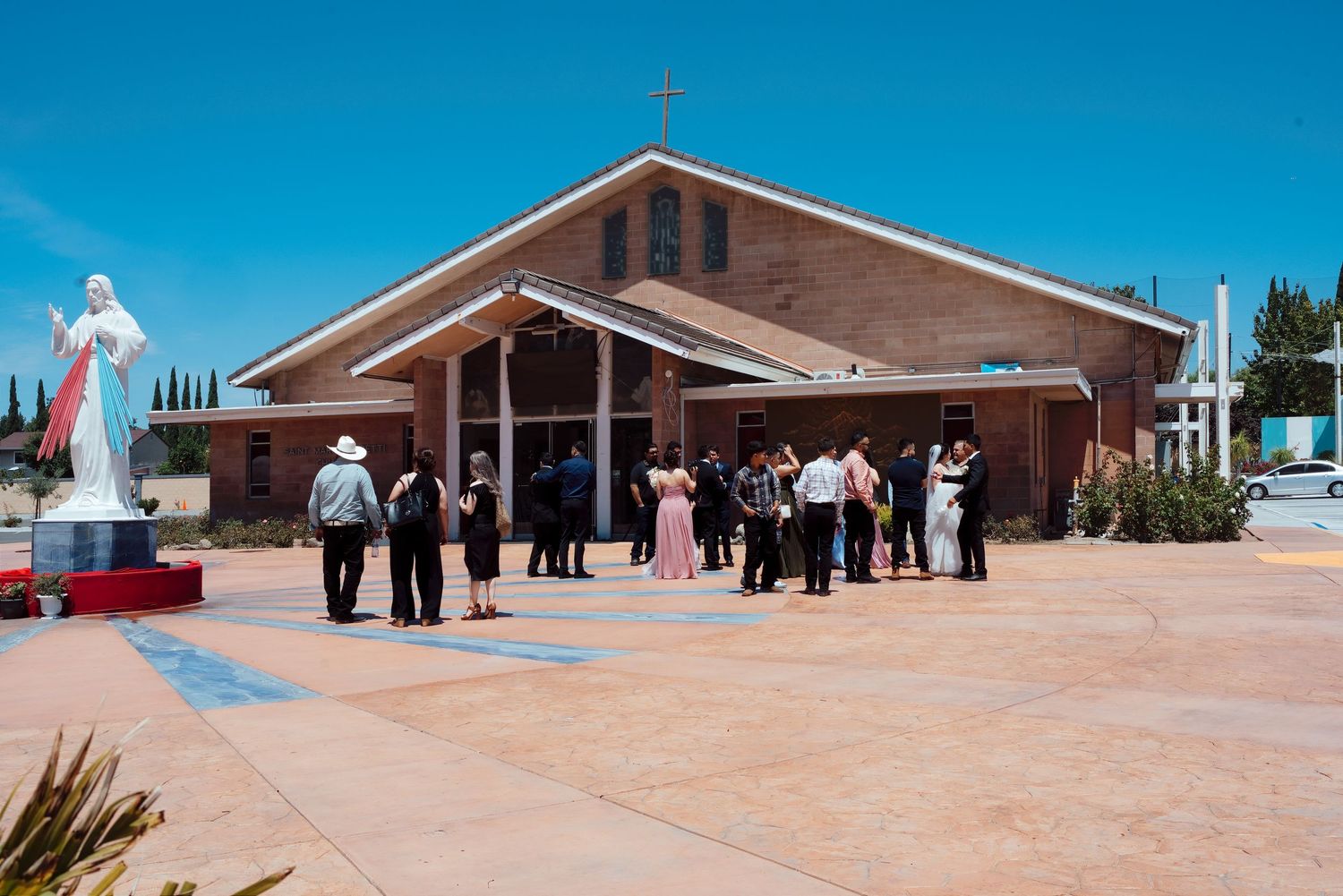 Guests gather outside a brick Catholic church with a Jesus statue under a bright blue sky.