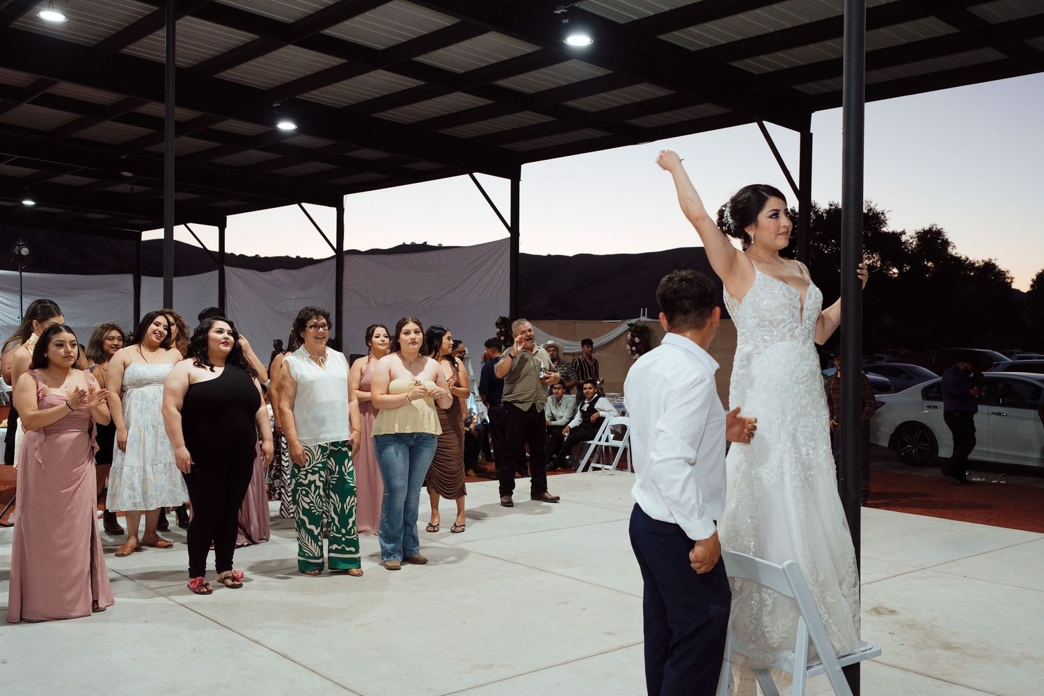 Bride tossing bouquet to excited wedding guests outdoors at evening reception under pavilion.