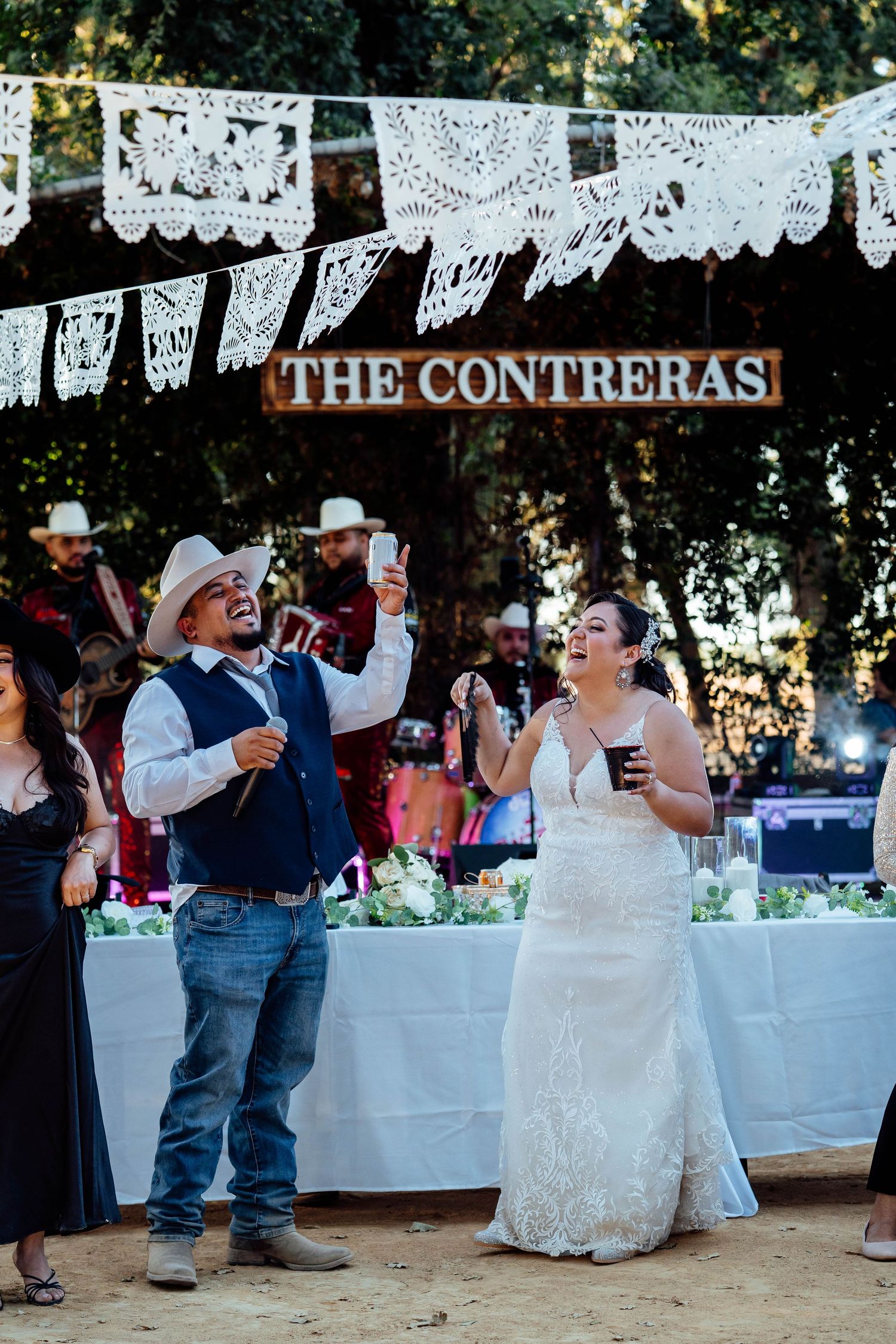 Bride and groom laugh and raise drinks under The Contreras sign at their outdoor wedding reception.
