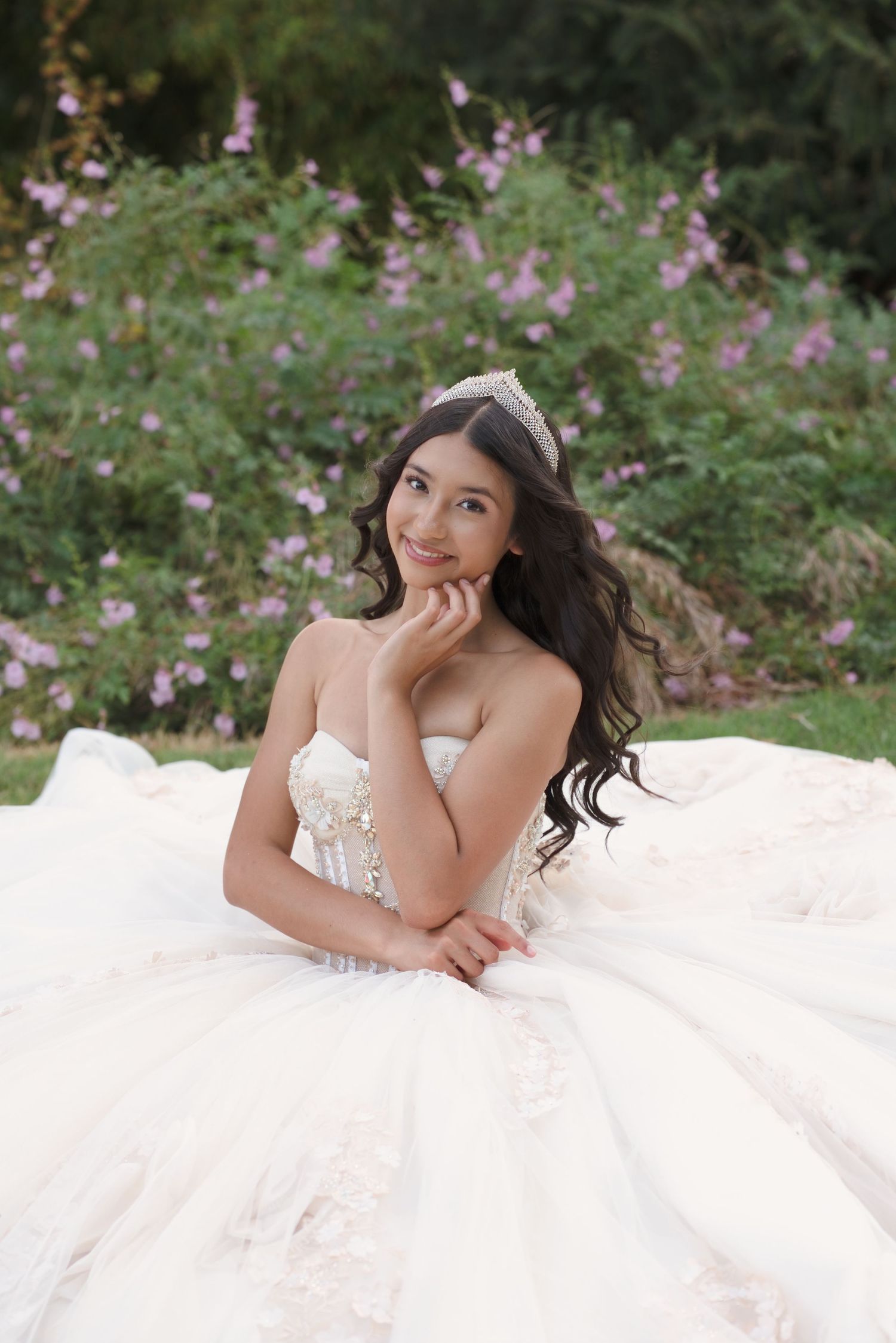 Young woman in a white ballgown and tiara sits gracefully outdoors among pink flowers for a quinceañera portrait.