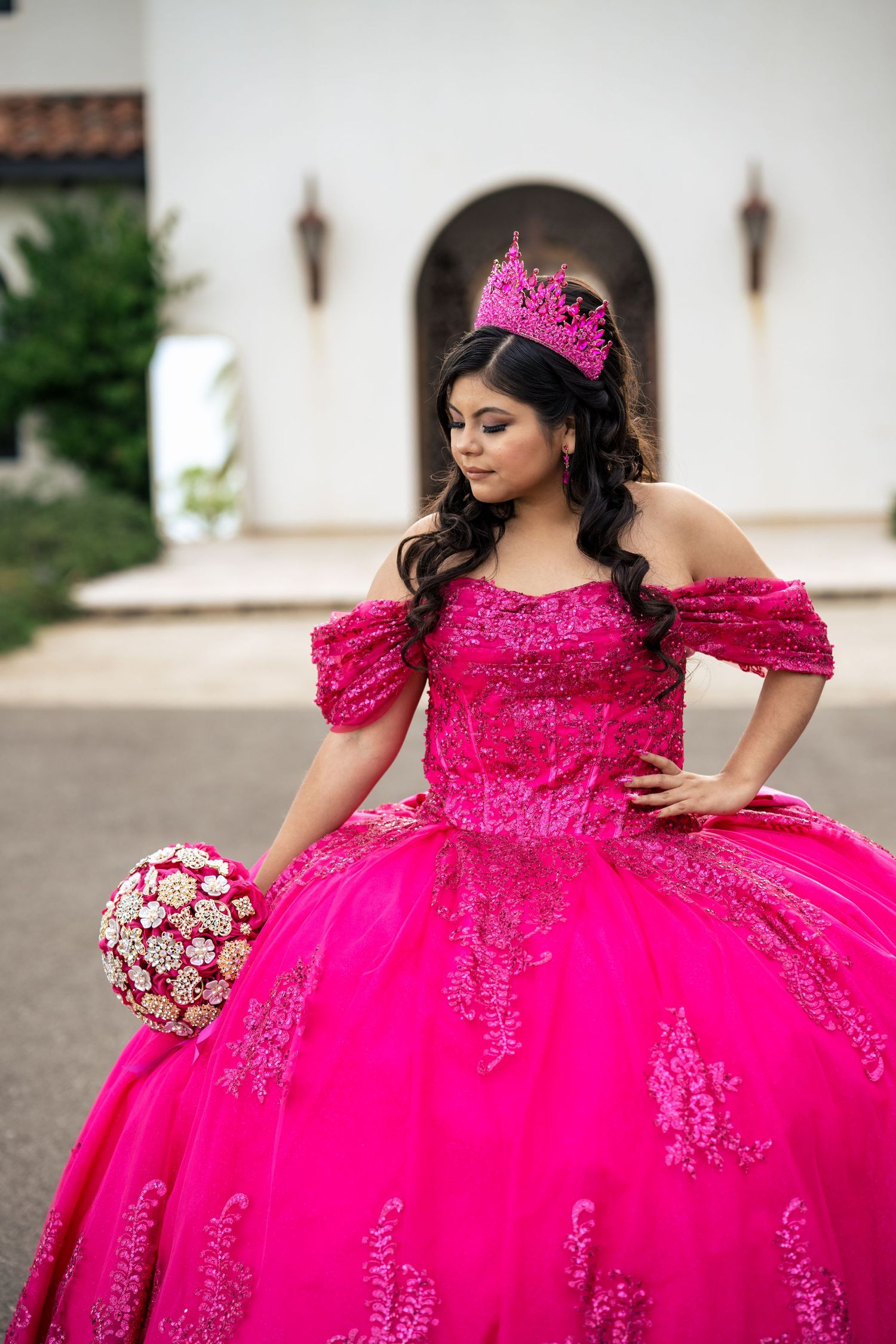 Young Latina in hot pink quinceañera ball gown with crown and floral bouquet poses outdoors.