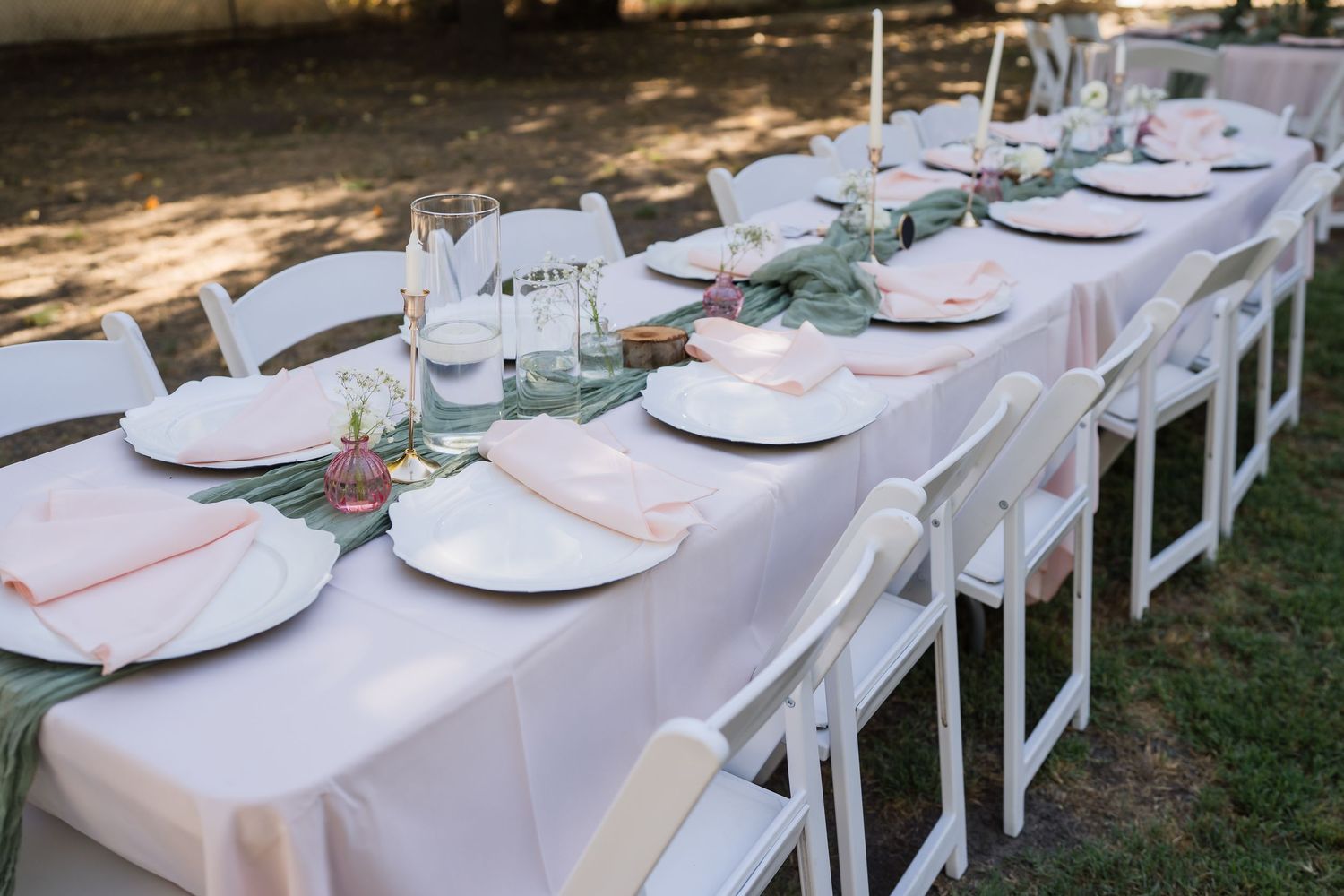 Elegant outdoor wedding reception table set with white chairs, pink napkins, sage greenery, and white plates on a blush tablecloth.