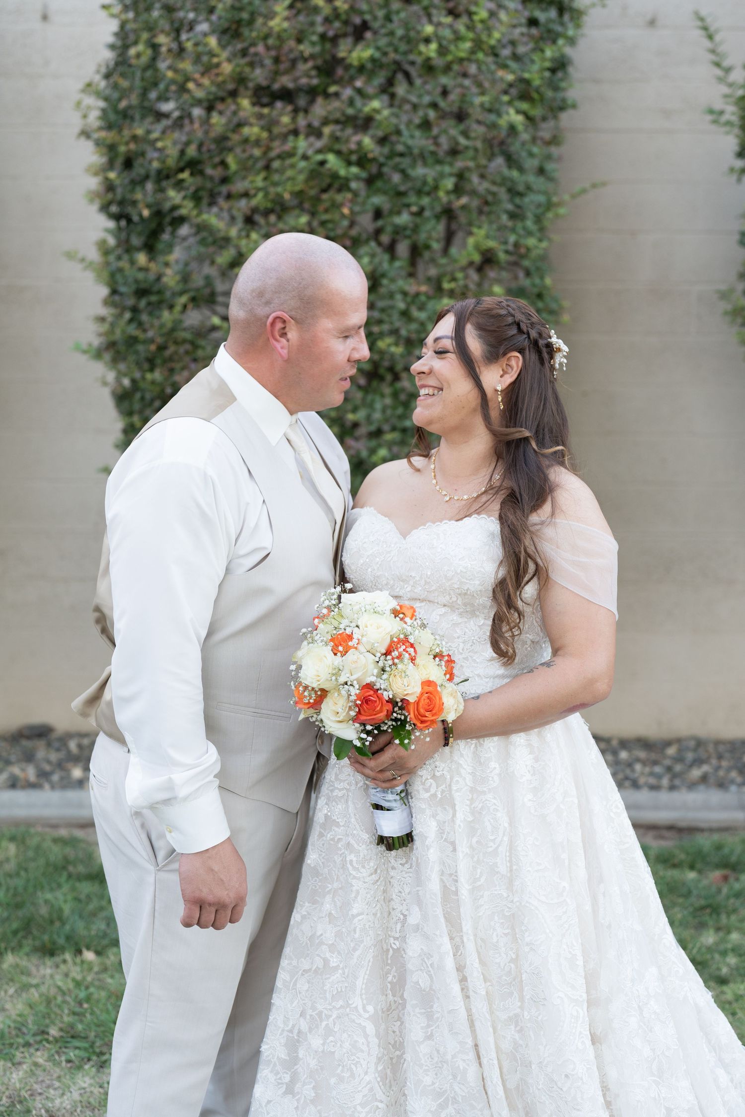 Bride and groom smiling at each other outdoors, bride holding orange and white bouquet, groom in tan suit.