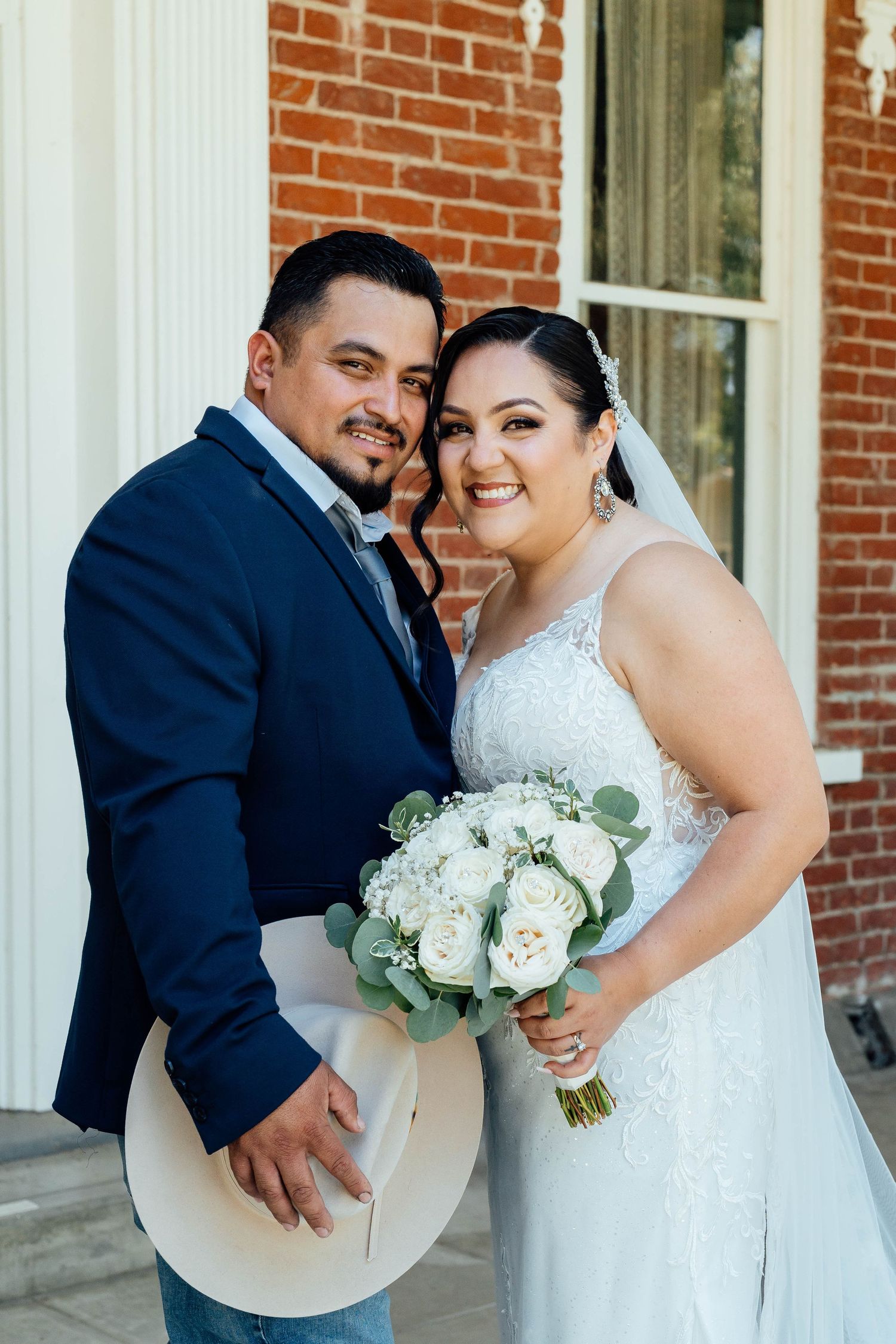 Happy bride and groom portrait outside brick building, bride holding white rose and eucalyptus bouquet.