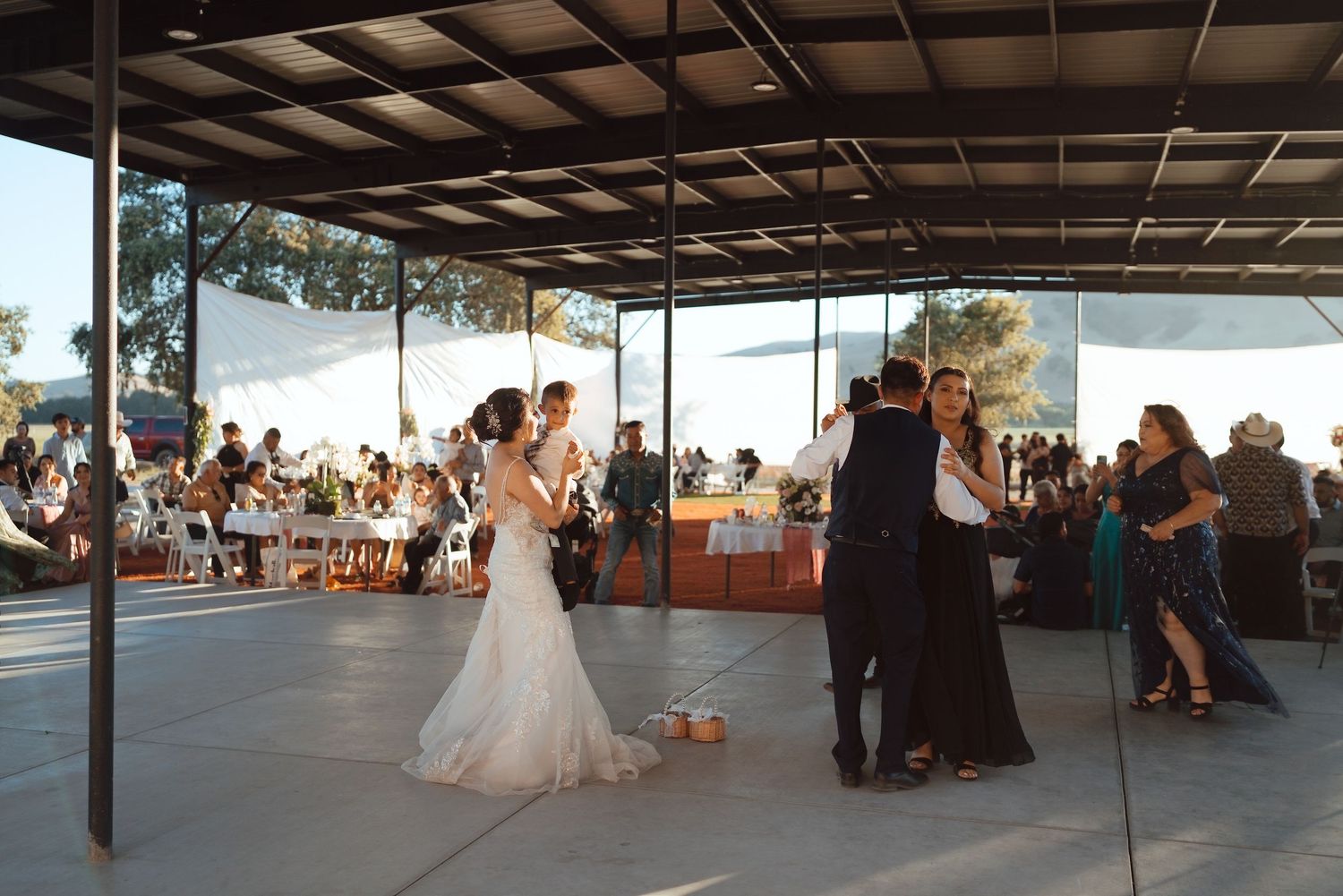 Couples share first dance at outdoor wedding reception under covered pavilion with guests seated at tables in background.