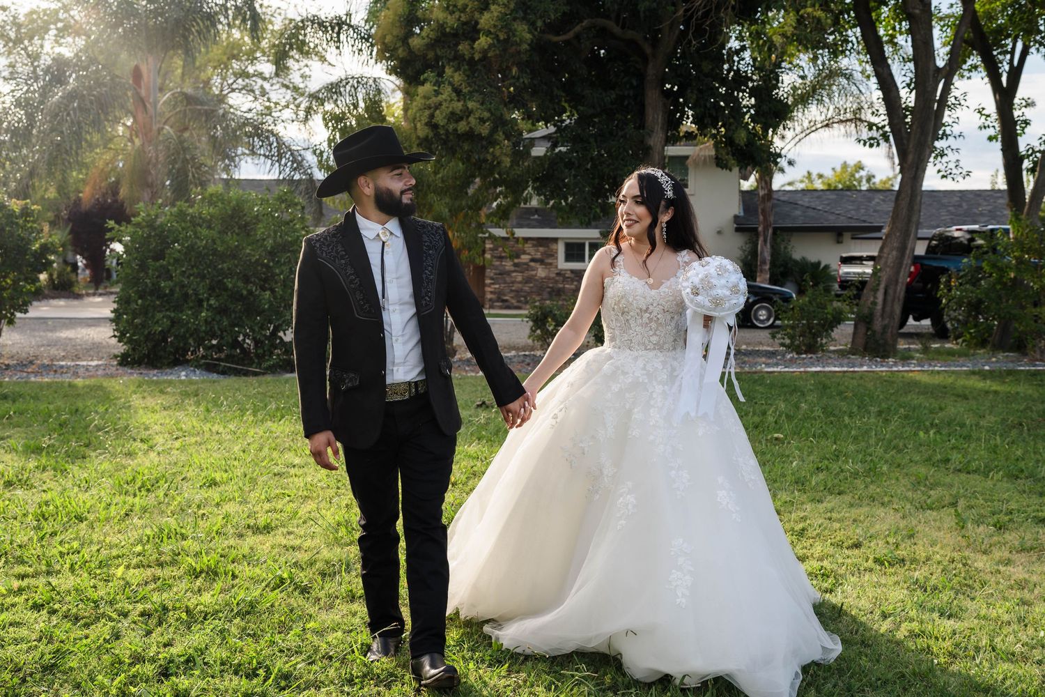 Bride in white ballgown and groom in black suit hold hands walking on green lawn in outdoor wedding photo.