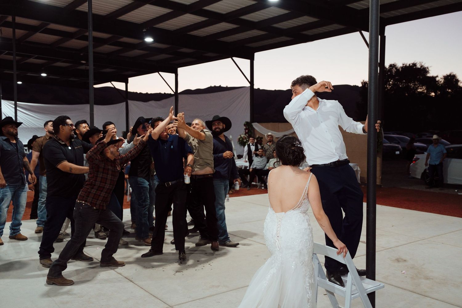 Bride and groom celebrate at outdoor wedding reception with guests cheering and dancing under a covered venue at dusk.