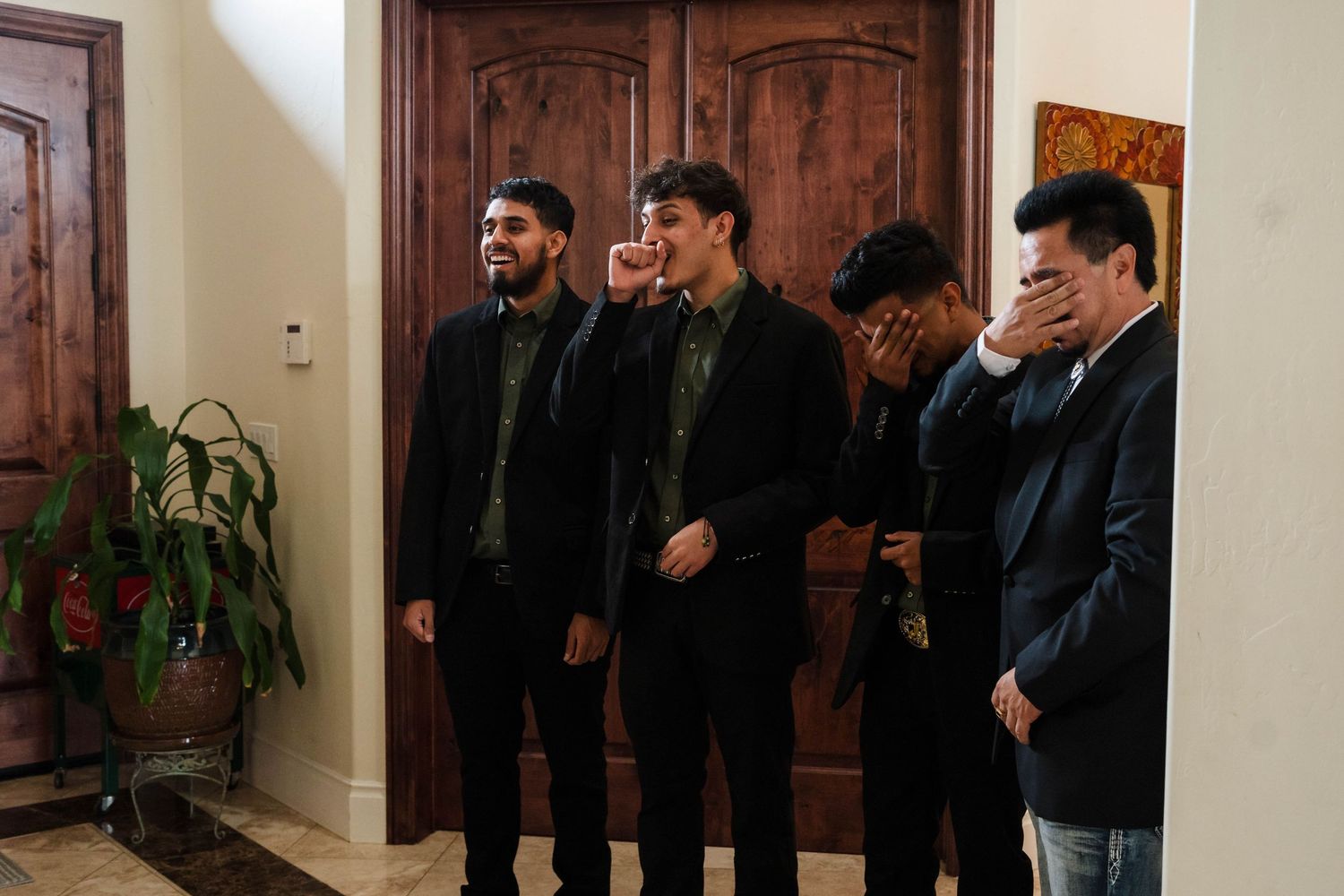 Four men in dark formal attire standing in an elegant foyer with wooden doors, some covering their faces emotionally.