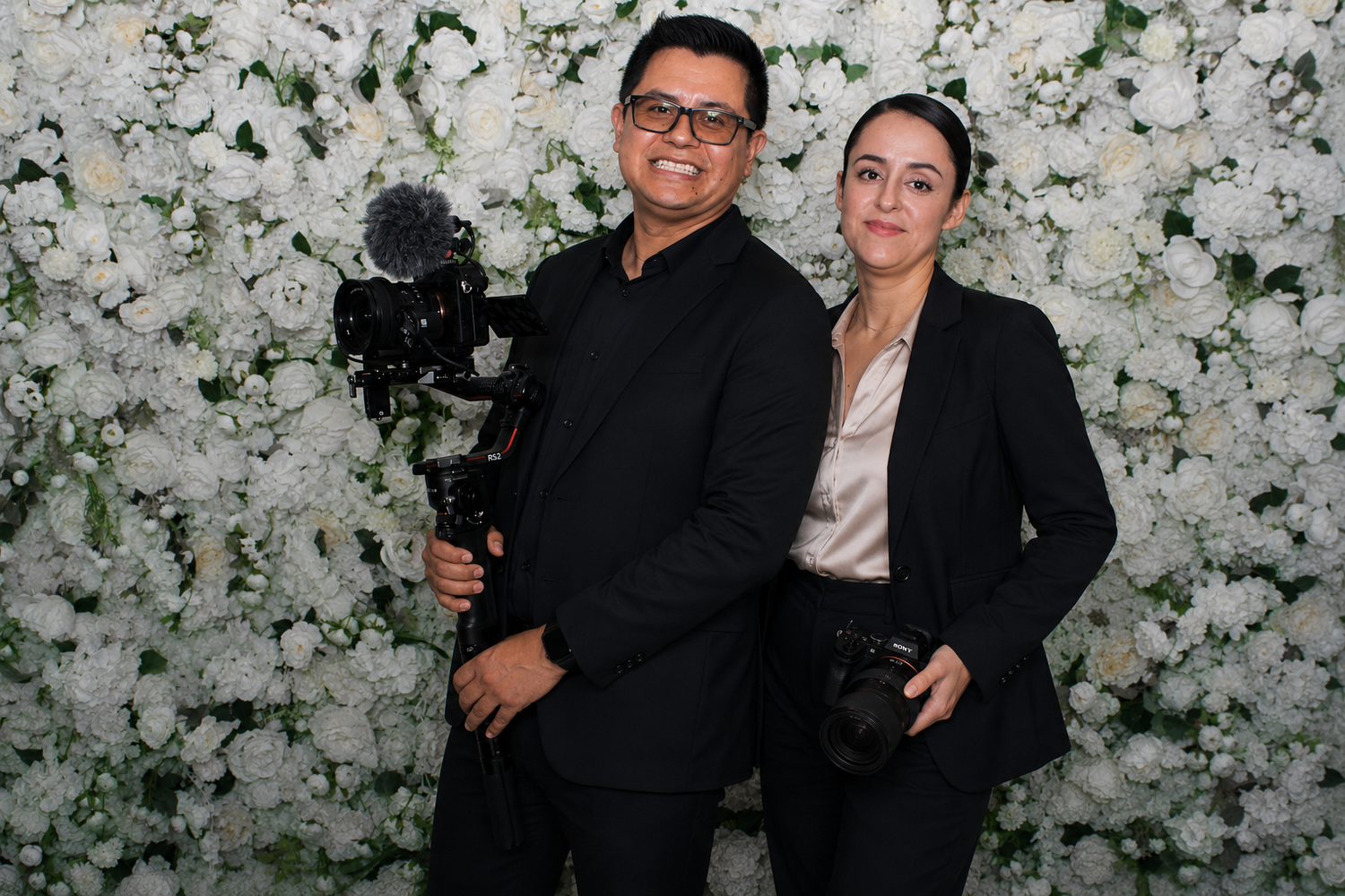 Two photographers pose with camera equipment in front of a white floral wall backdrop at an event.