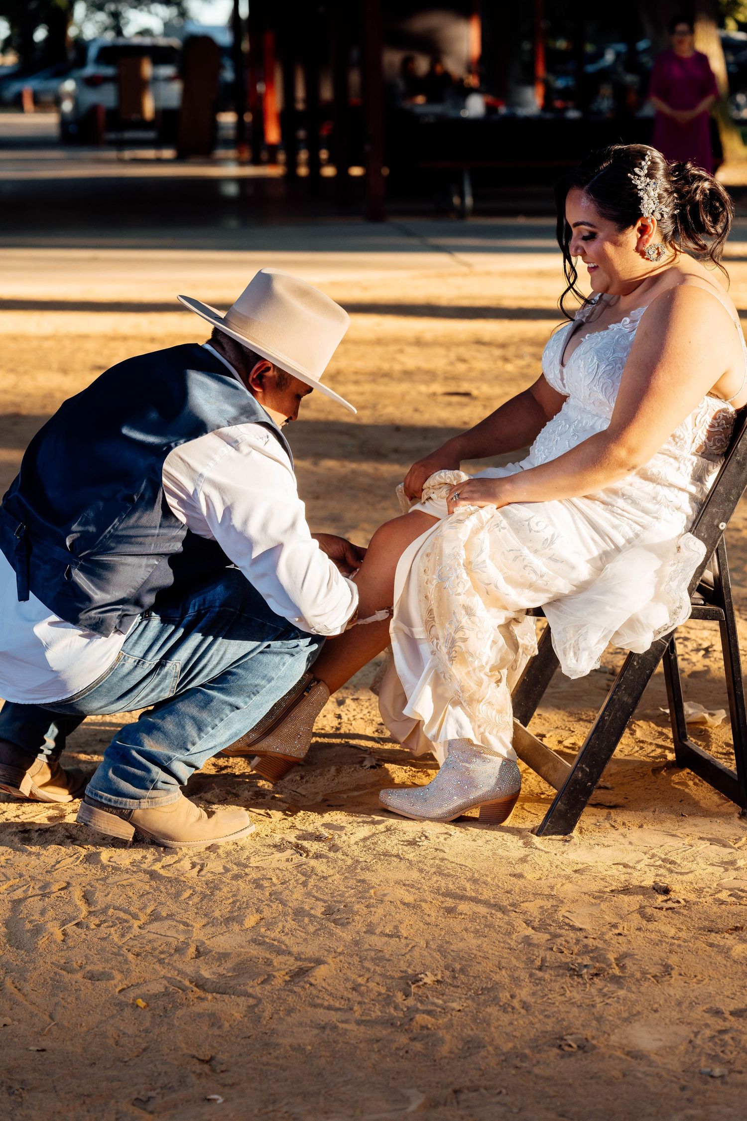 Groom in cowboy hat kneeling to adjust bride's shoe during outdoor wedding ceremony on sandy ground.