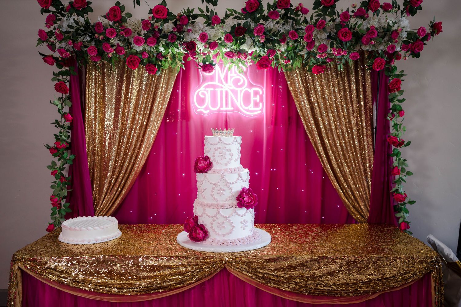 Elegant three-tier white quinceañera cake displayed under a glowing neon sign with floral backdrop.
