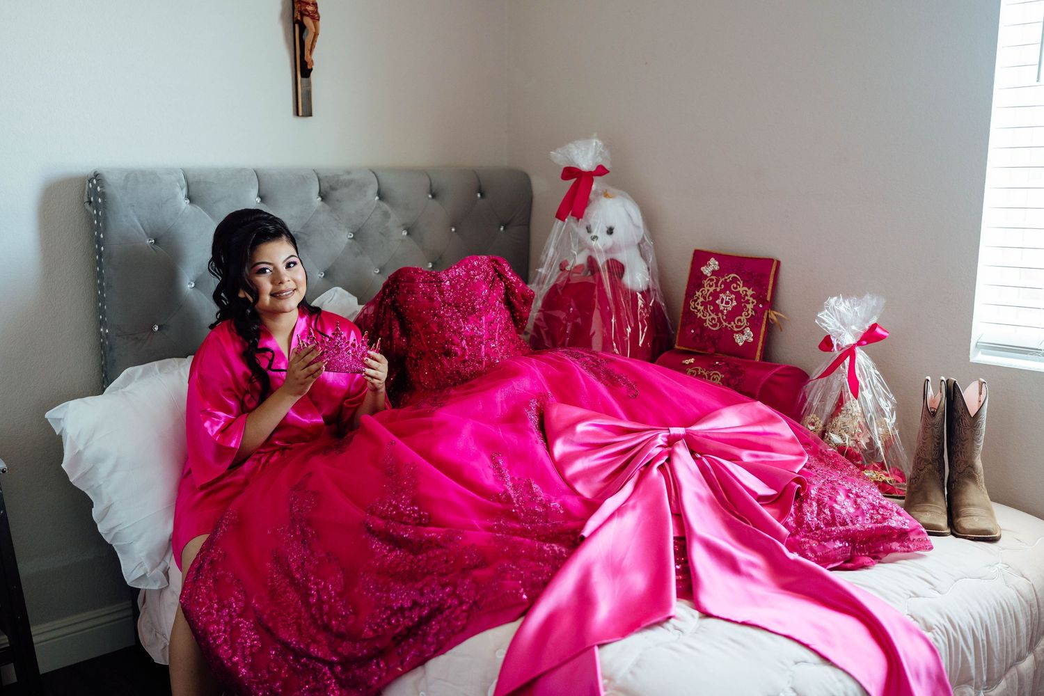 Quinceañera girl in hot pink ball gown sitting on bed surrounded by gifts and decorations.