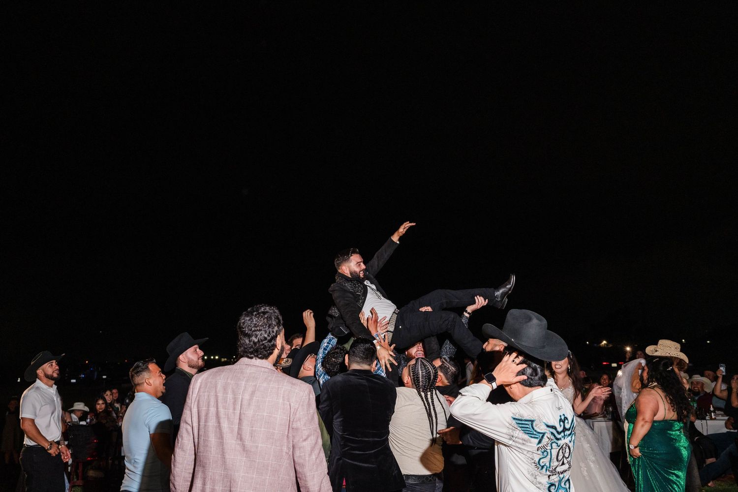 Wedding guests toss the groom into the air during a lively outdoor nighttime wedding reception celebration.