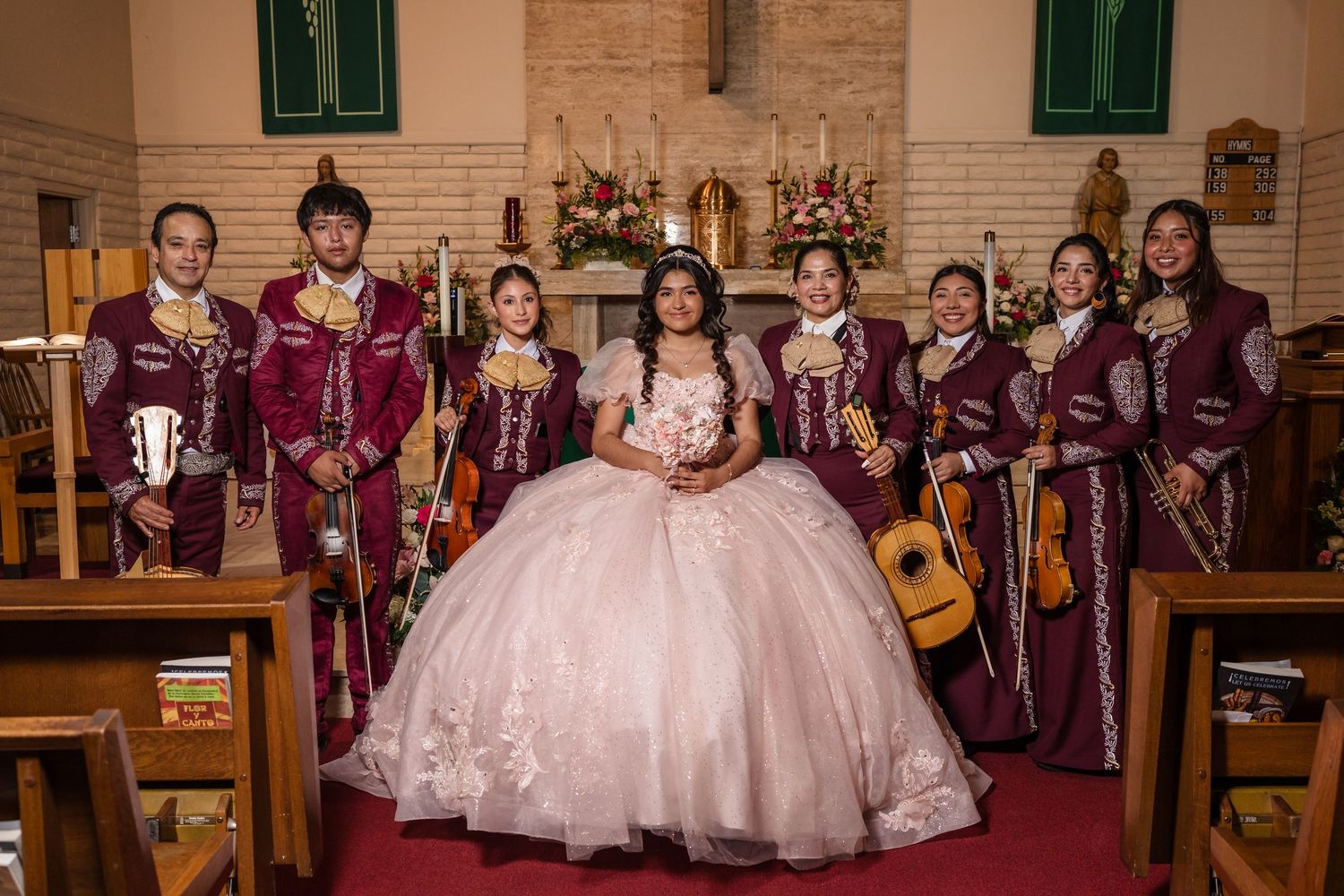 Young woman in pink quinceañera gown poses with mariachi band in matching burgundy suits inside a church.