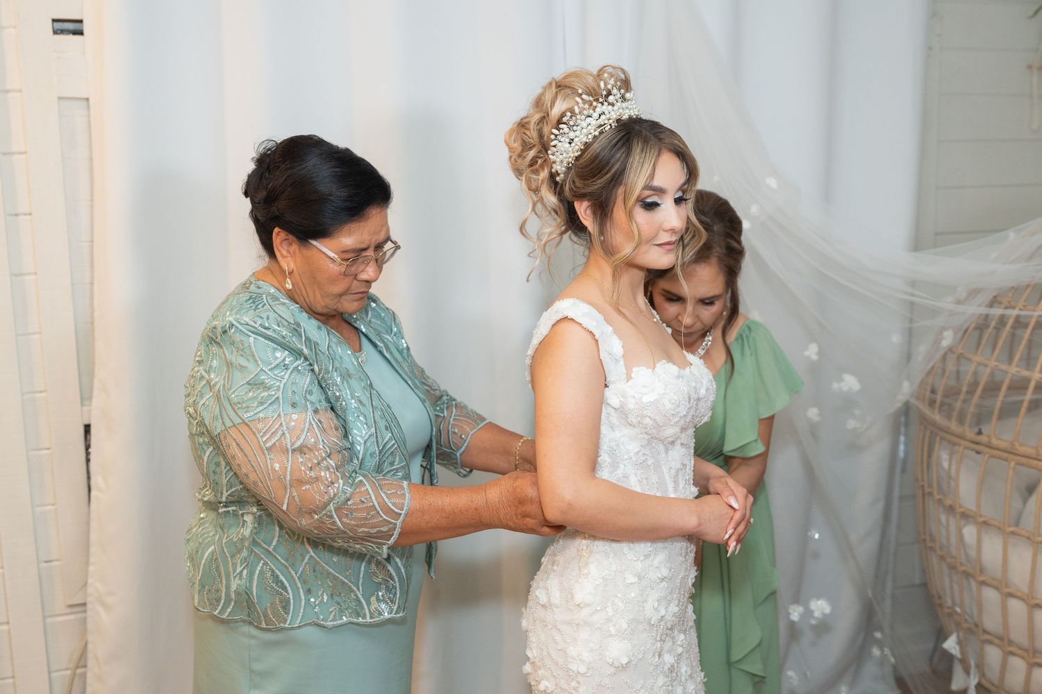 Bride in white lace gown being helped by two women, one in teal, during wedding preparations indoors.