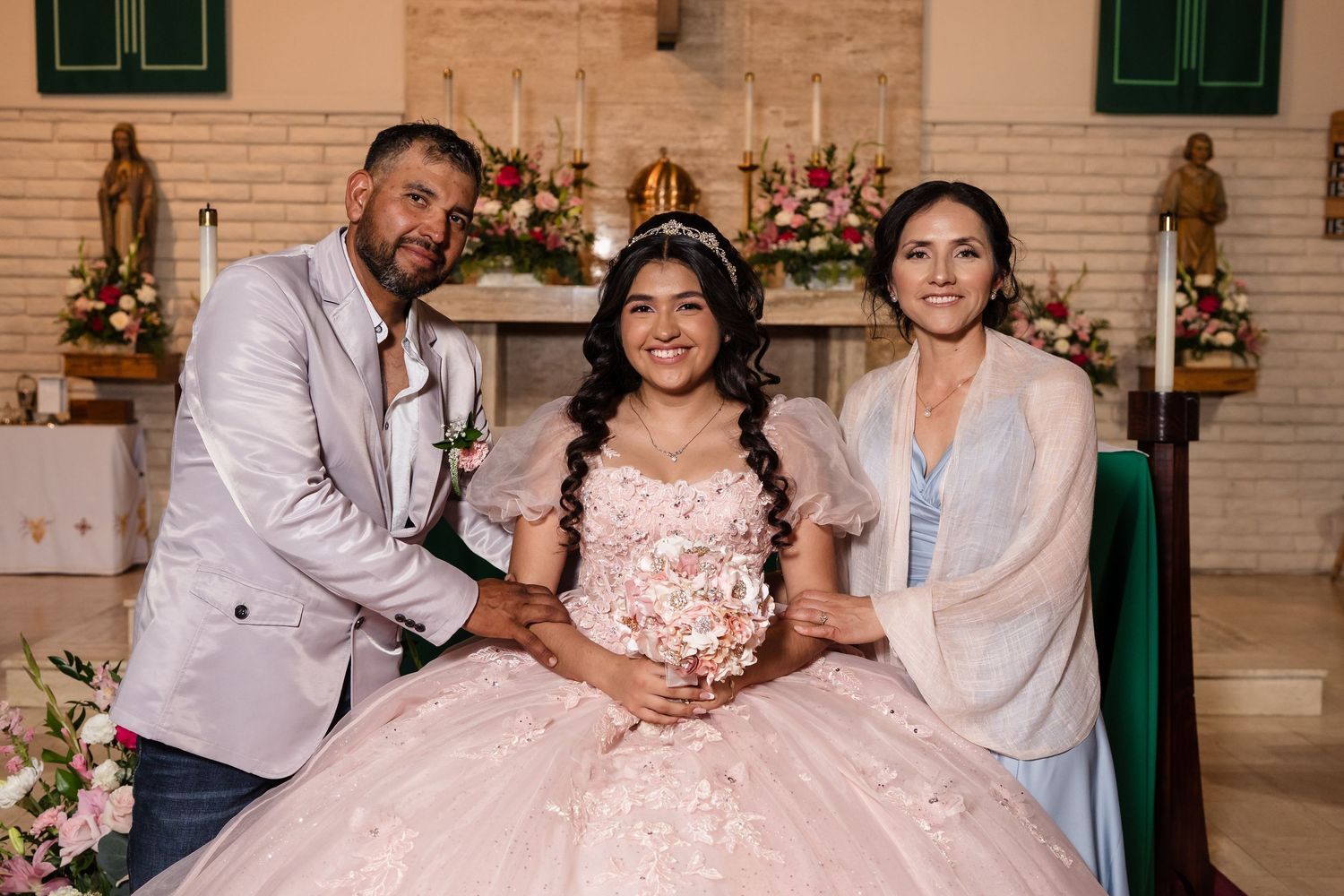 A young girl in a pink quinceañera gown poses with her parents inside a decorated church.