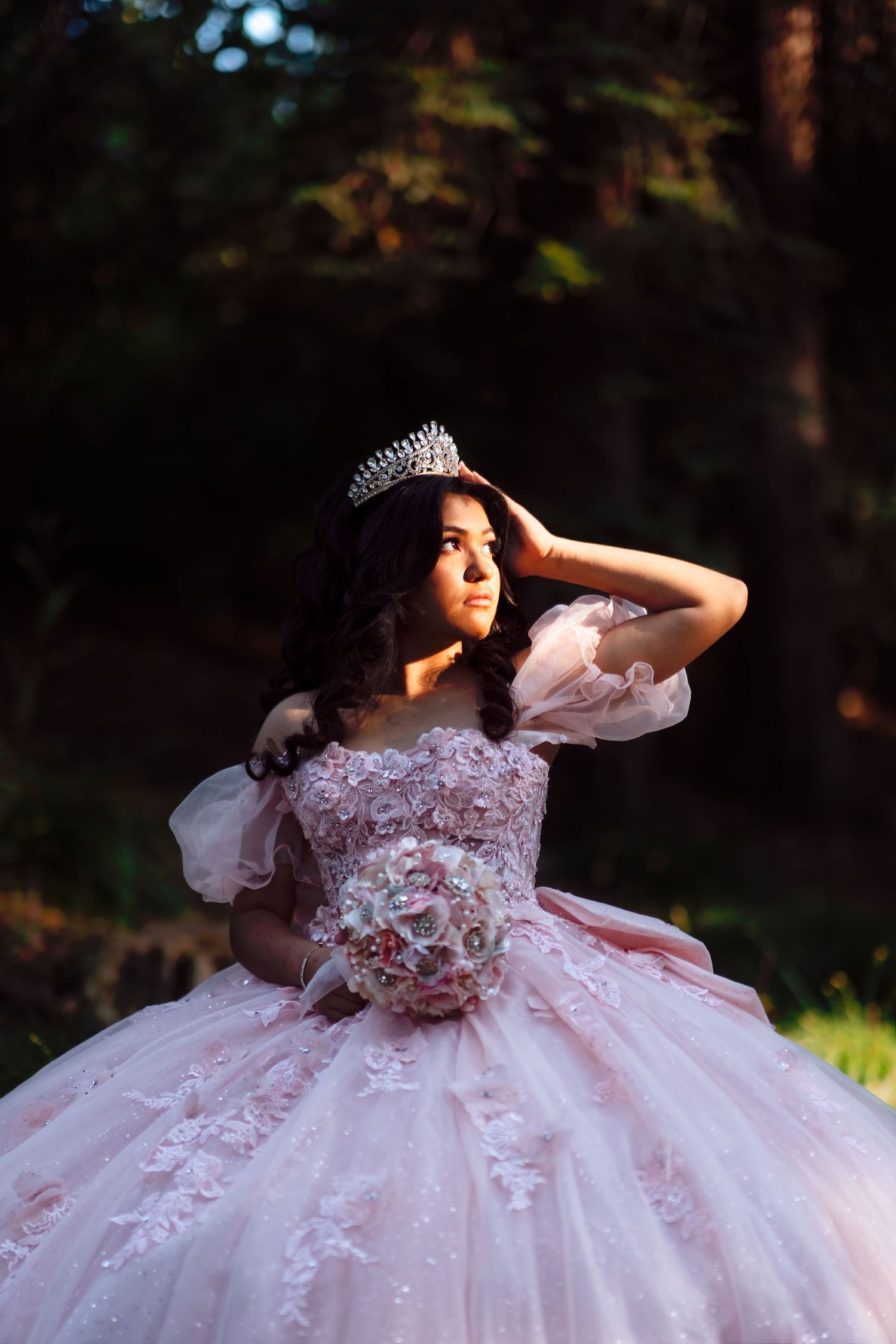 Young woman in a pink quinceañera ball gown wearing a tiara, holding a bouquet in a sunlit forest.