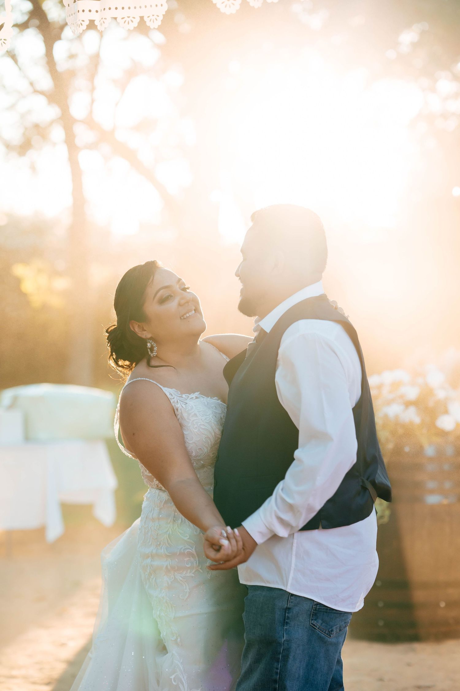 Bride and groom holding hands and smiling at each other in golden sunset light at wedding.