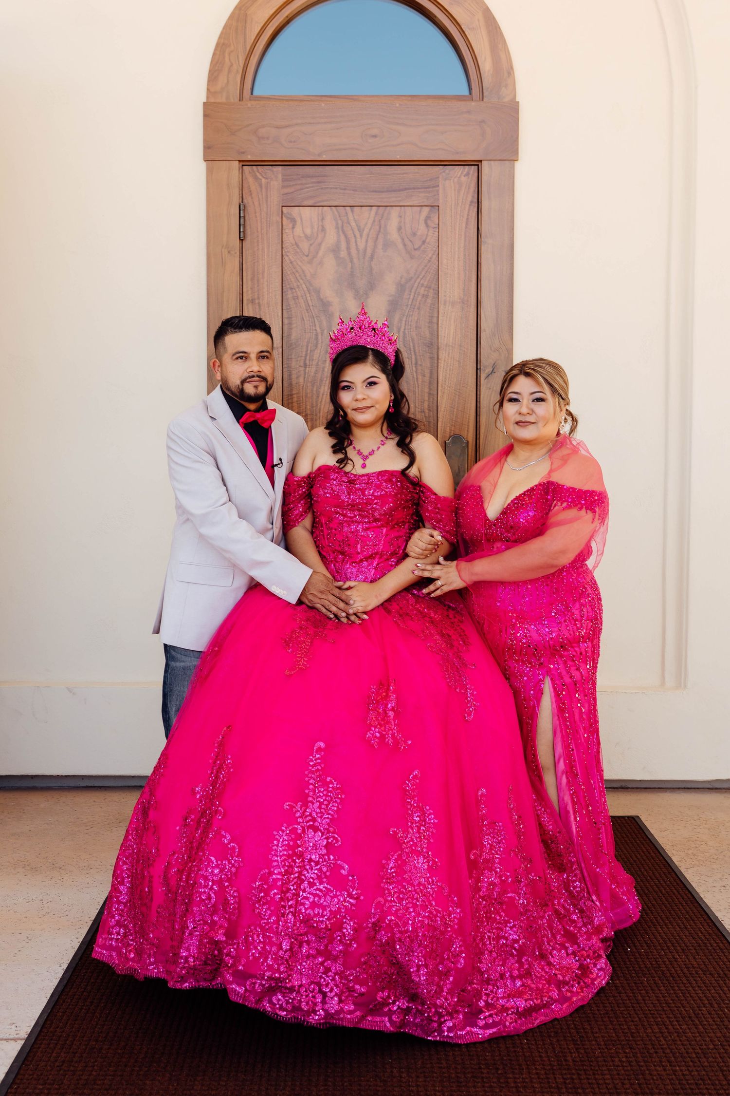 Quinceañera girl in hot pink gown and crown posing with parents in front of wooden door.