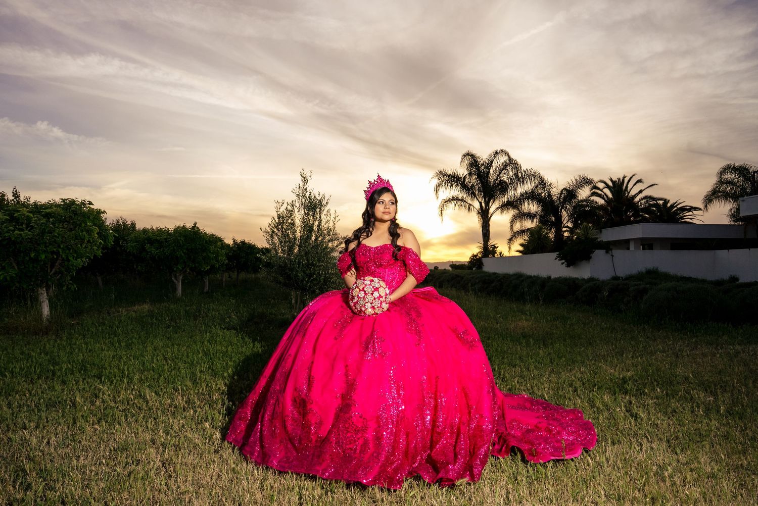 Quinceañera girl in a voluminous hot pink ball gown poses outdoors at sunset with palm trees behind her.