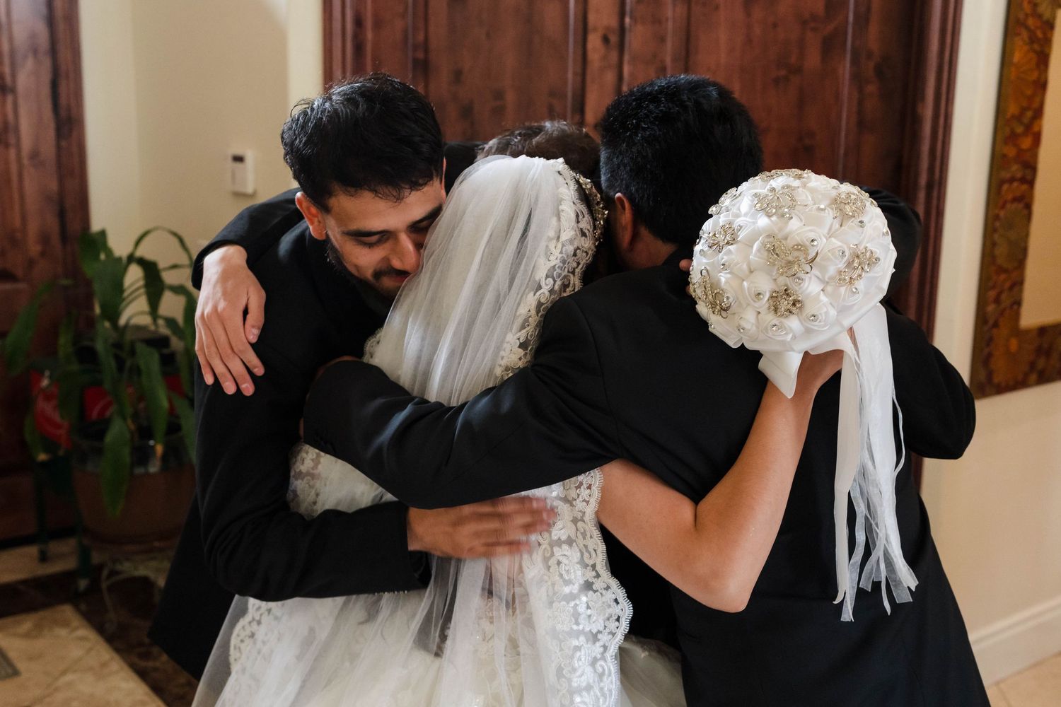 Bride in white lace gown and veil shares a group hug with two guests in black suits holding a bouquet.