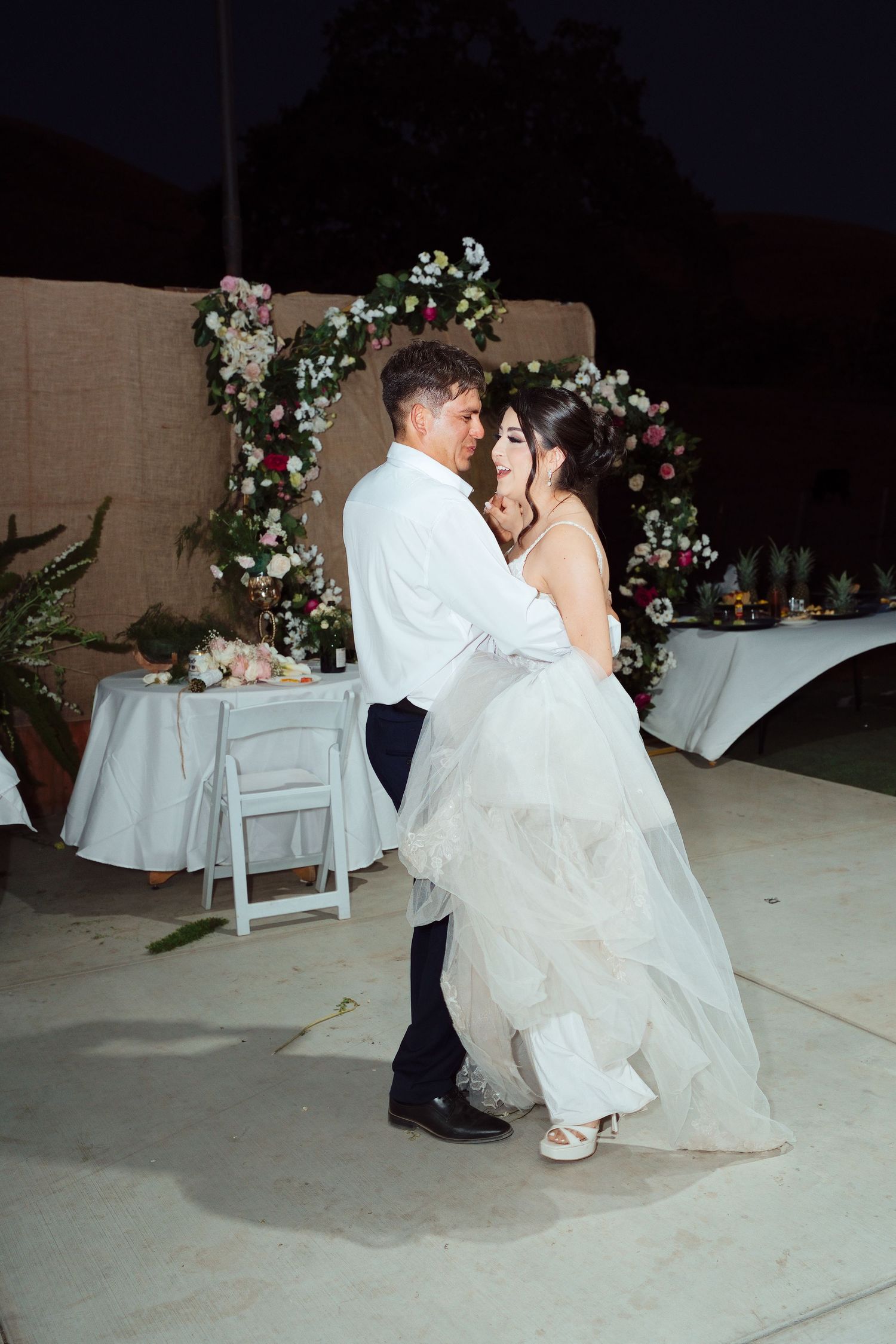 Bride and groom share their first dance outdoors at night, with a floral arch backdrop at their wedding reception.