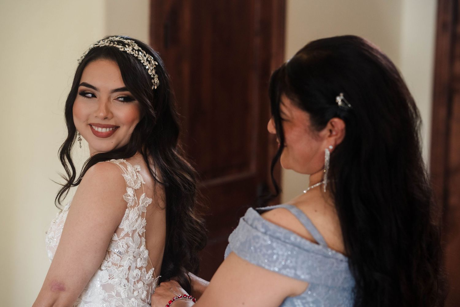 Bride in white lace gown and tiara smiles as woman in blue dress helps her prepare for wedding ceremony.