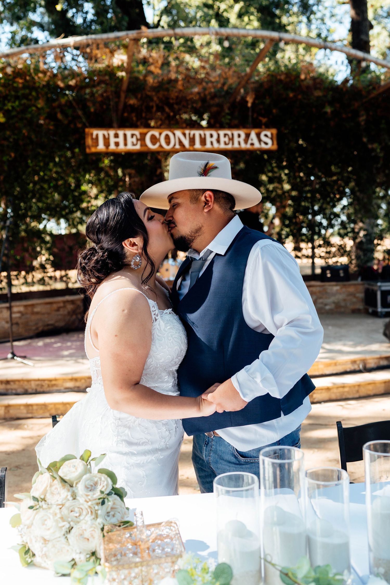 Newlyweds kiss at reception table under The Contreras sign with white flowers.