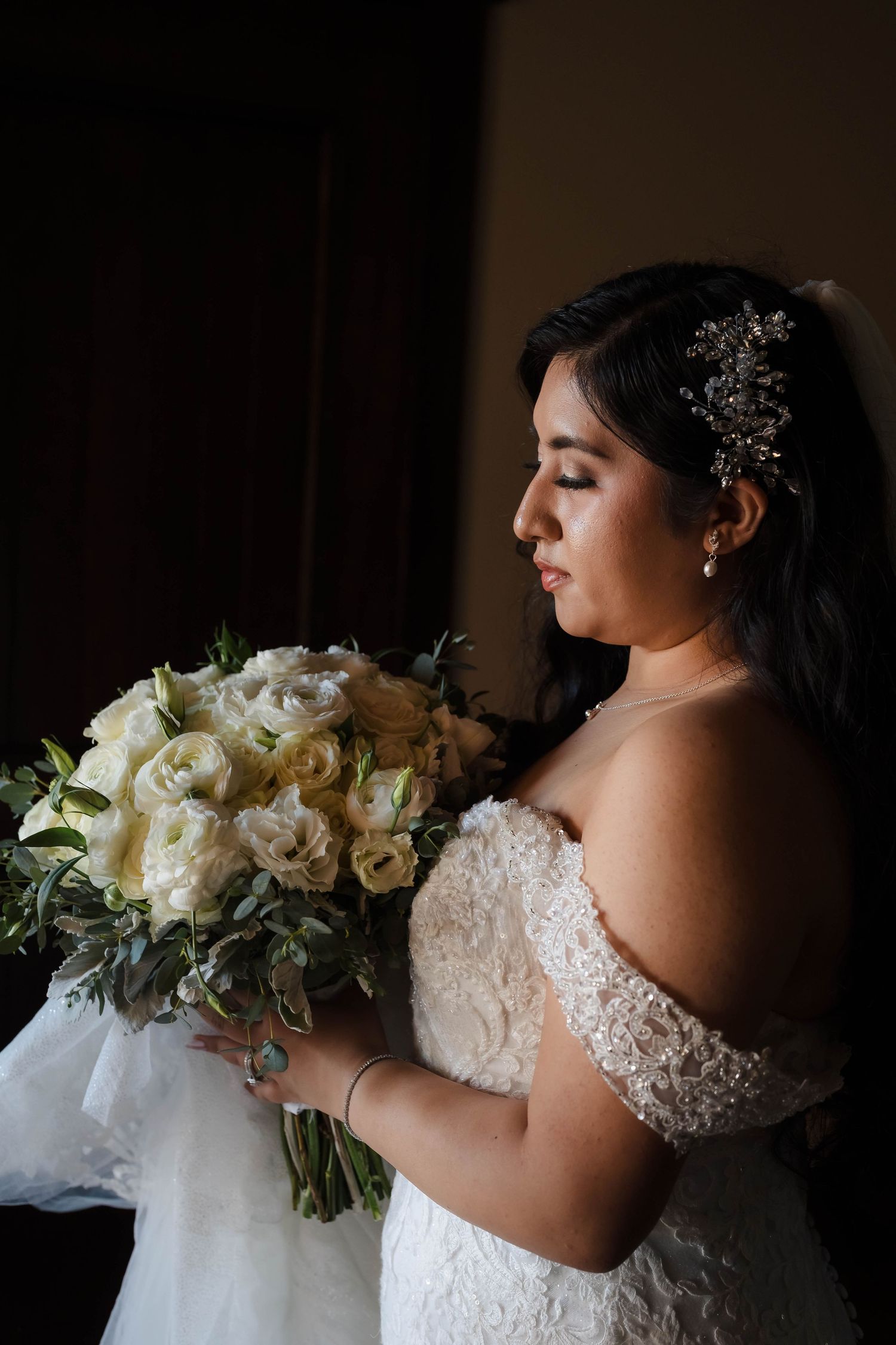 Bride in off-shoulder lace wedding gown holding a white rose bouquet, gazing down by a dark window.