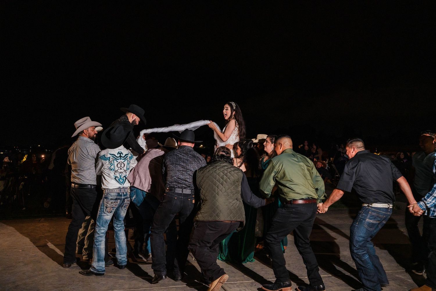 Group of people lifting a woman at an outdoor night celebration, surrounded by cheering guests.