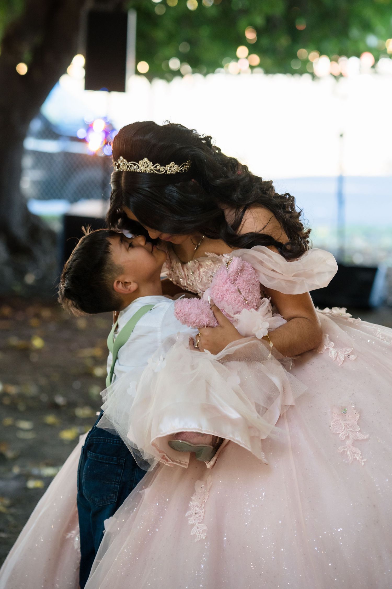 A quinceañera in a pink ball gown kisses a young boy in formal attire during her celebration outdoors.