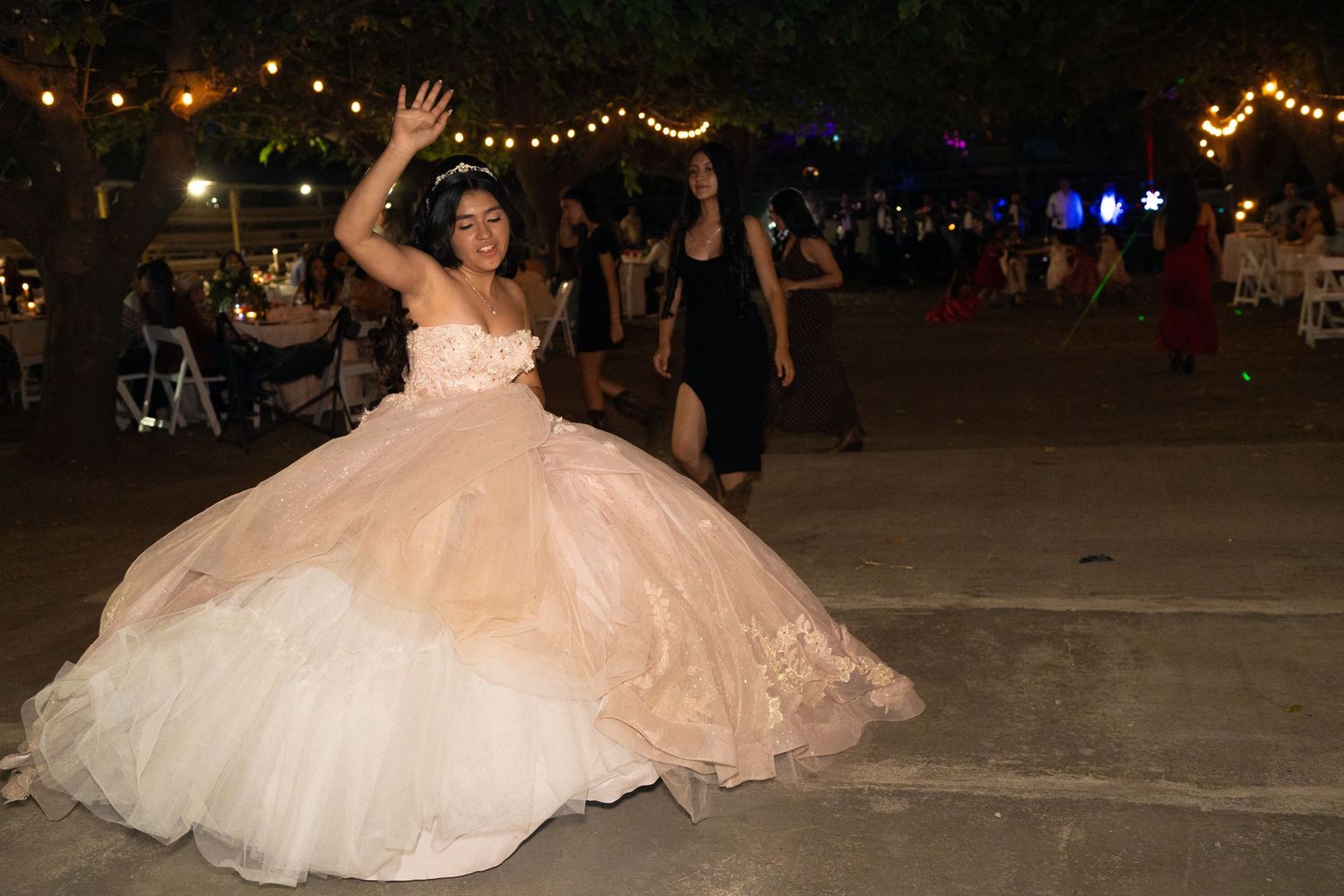 A quinceañera dances joyfully in a blush pink ball gown under string lights at her outdoor party.