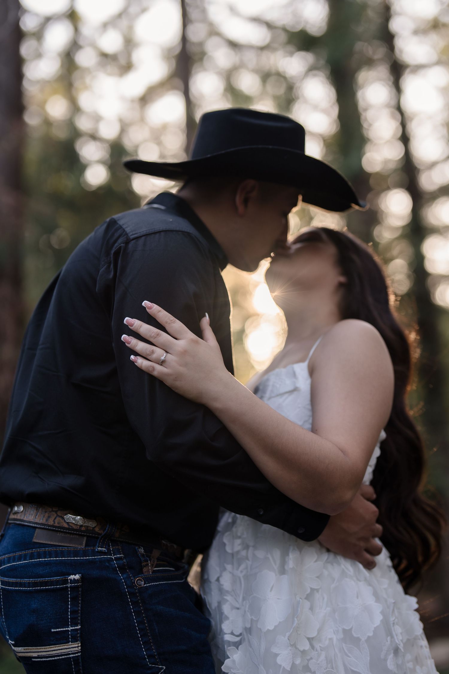 Couple sharing a romantic kiss in a forest, man wearing black cowboy hat and shirt, woman in white lace dress, engagement ring visible.