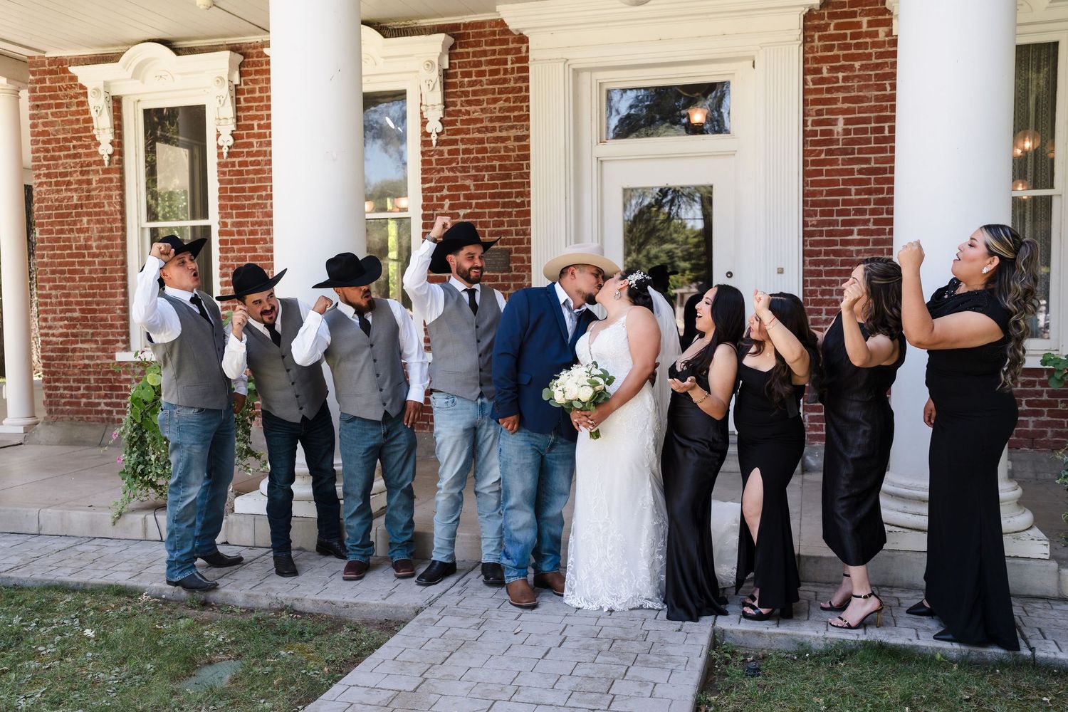 Wedding party poses outside brick mansion, groomsmen in cowboy hats, bridesmaids in black, bride and groom kissing.